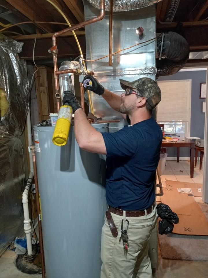Man working on HVAC system, wearing safety glasses and gloves, in a basement with pipes and ductwork.