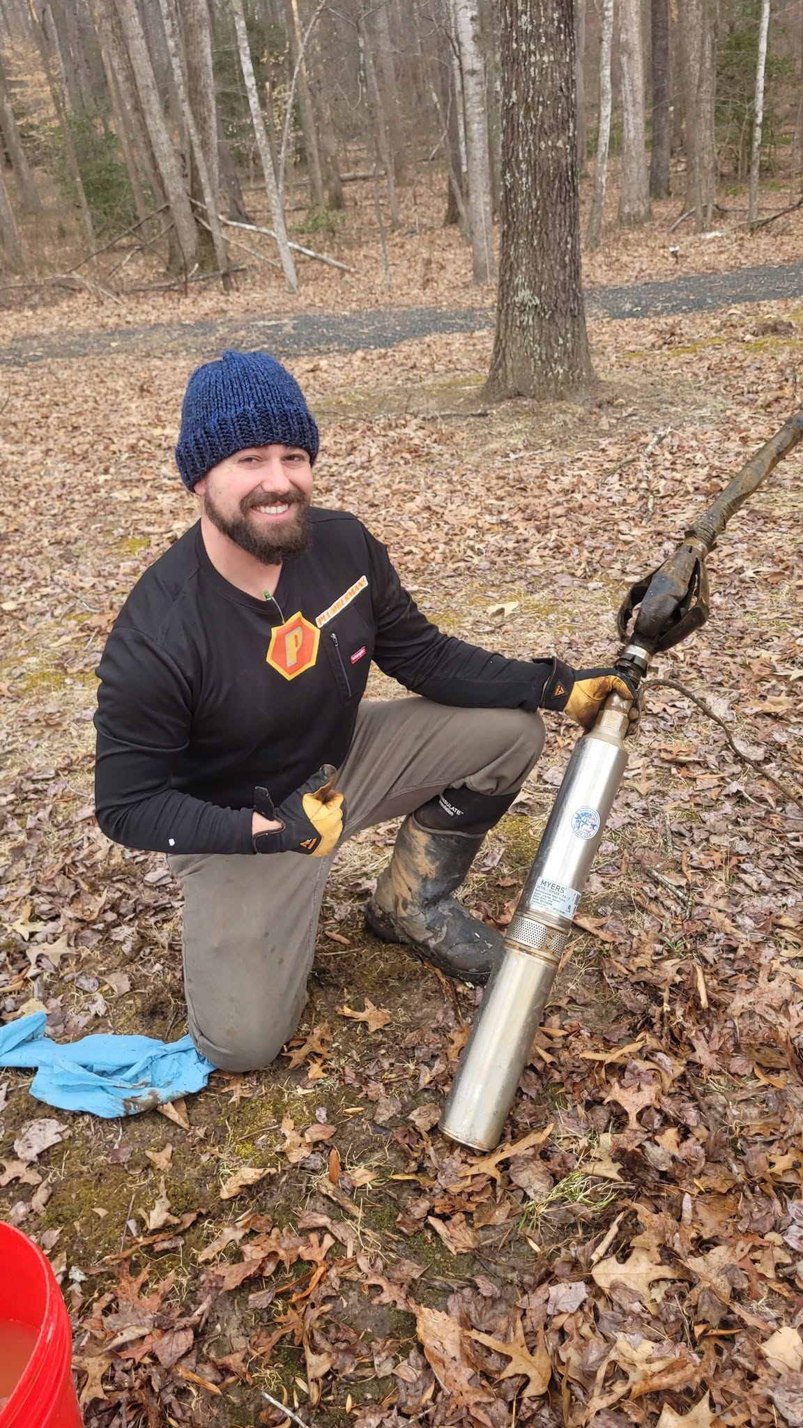 Person kneeling outdoors in a forest, wearing a blue knit hat and black jacket, with a large cylindrical device and a stick.
