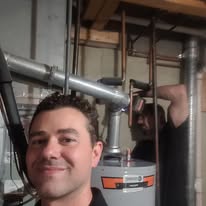 Man smiling in a workshop with pipes and tools in the background.
