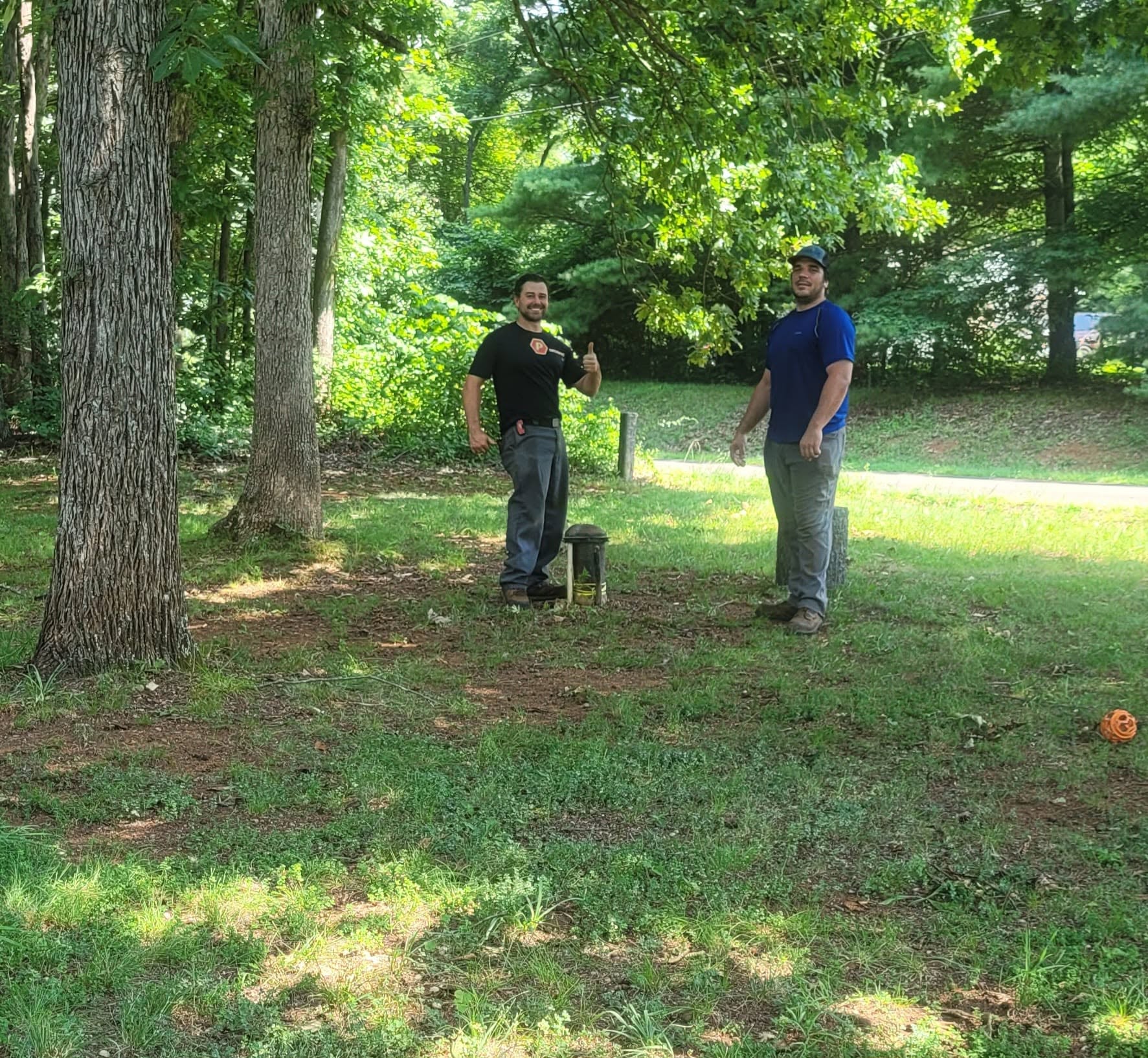 Two men stand outdoors in a wooded area, talking, with a small black dog between them, and a small orange ball on the ground.