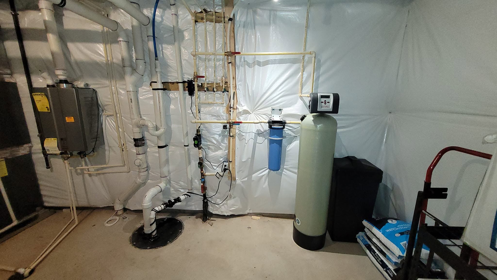 Indoor utility room with pipes, a water filter, and equipment against a white wall.