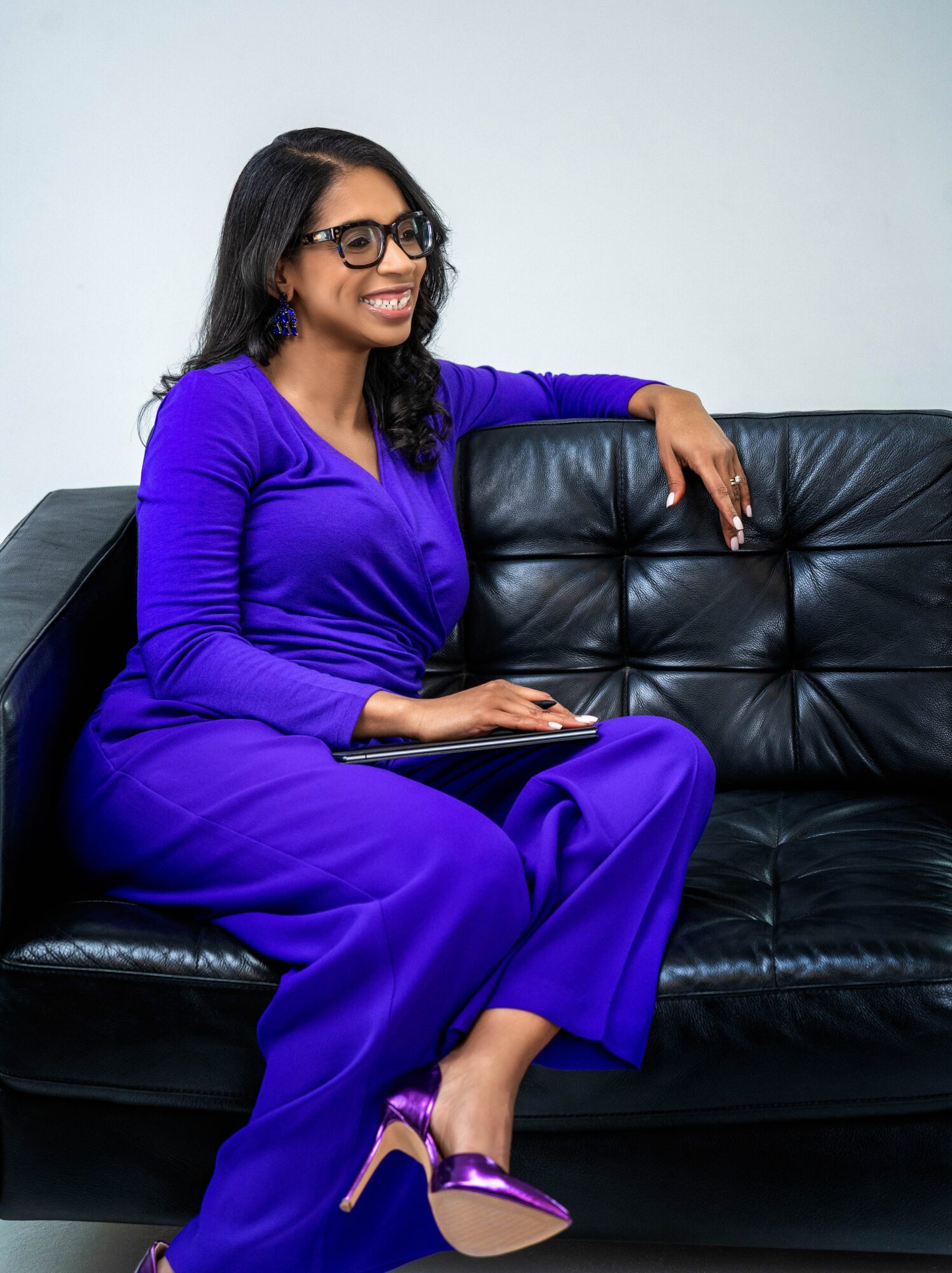 Woman in purple dress and glasses sitting on a black leather couch, smiling, holding a tablet, with a light background.