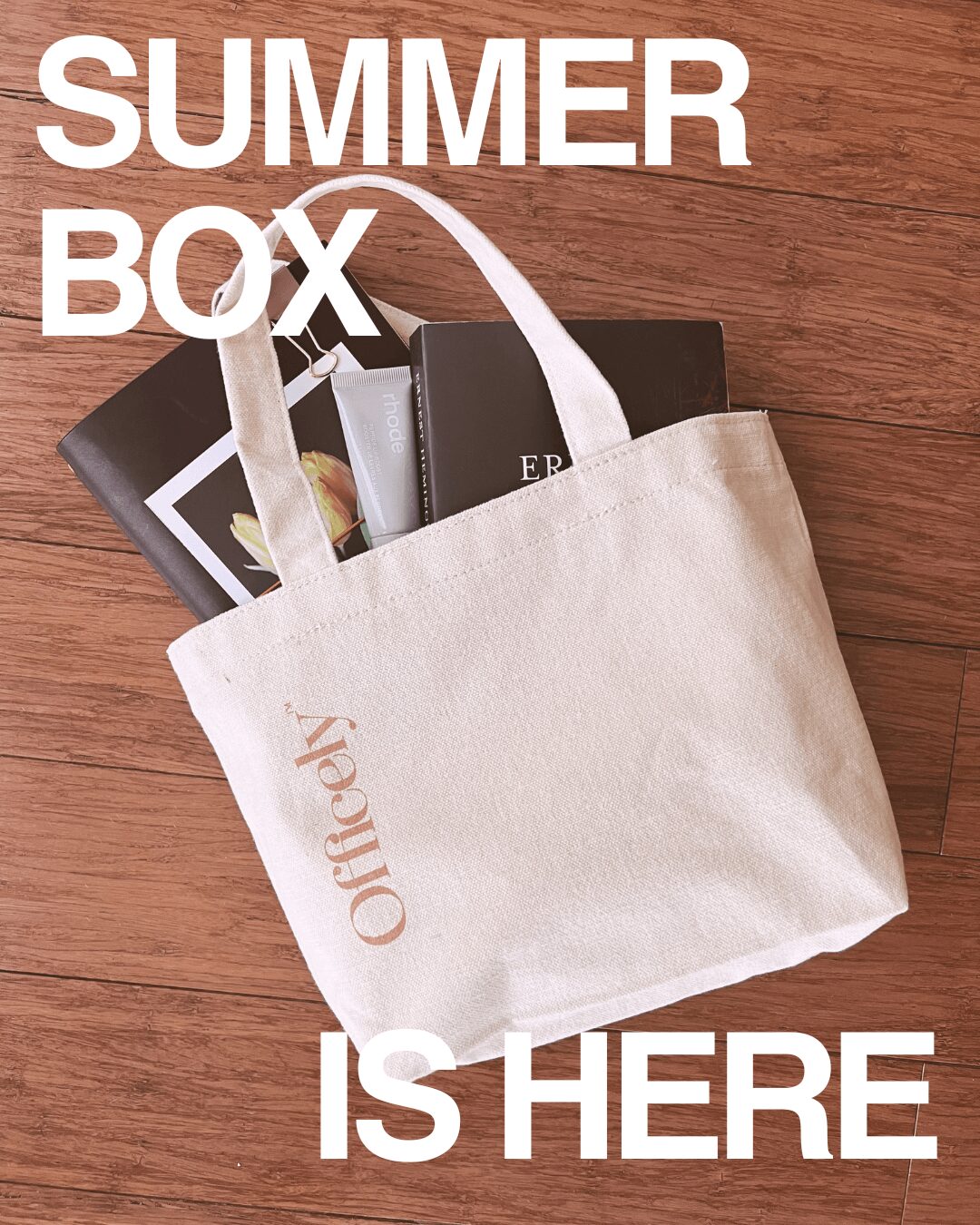White tote bag with items inside, on wooden surface, with text 'SUMMER BOX IS HERE' above and below, 20 words.