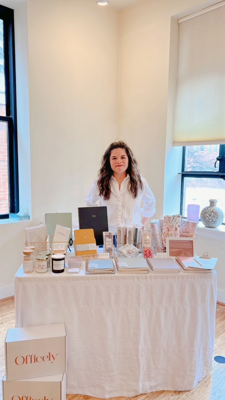 Woman sitting behind a table with various products, in a bright room with large windows and a white wall.