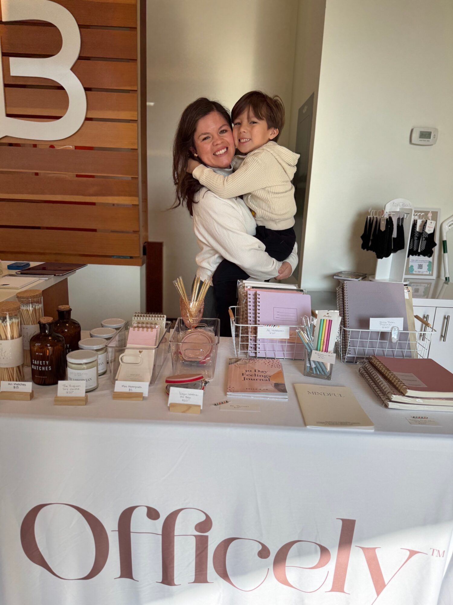 Two children hugging and smiling behind a reception desk in an office setting, with office supplies and a wooden wall in the background.