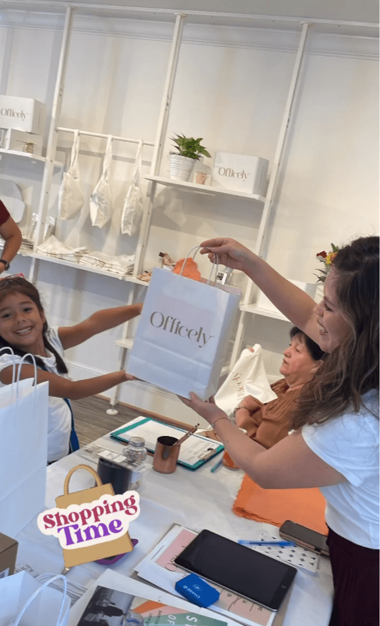 Child receiving a gift from an adult at a table with shopping items, smiling girl, store background, bright lighting.