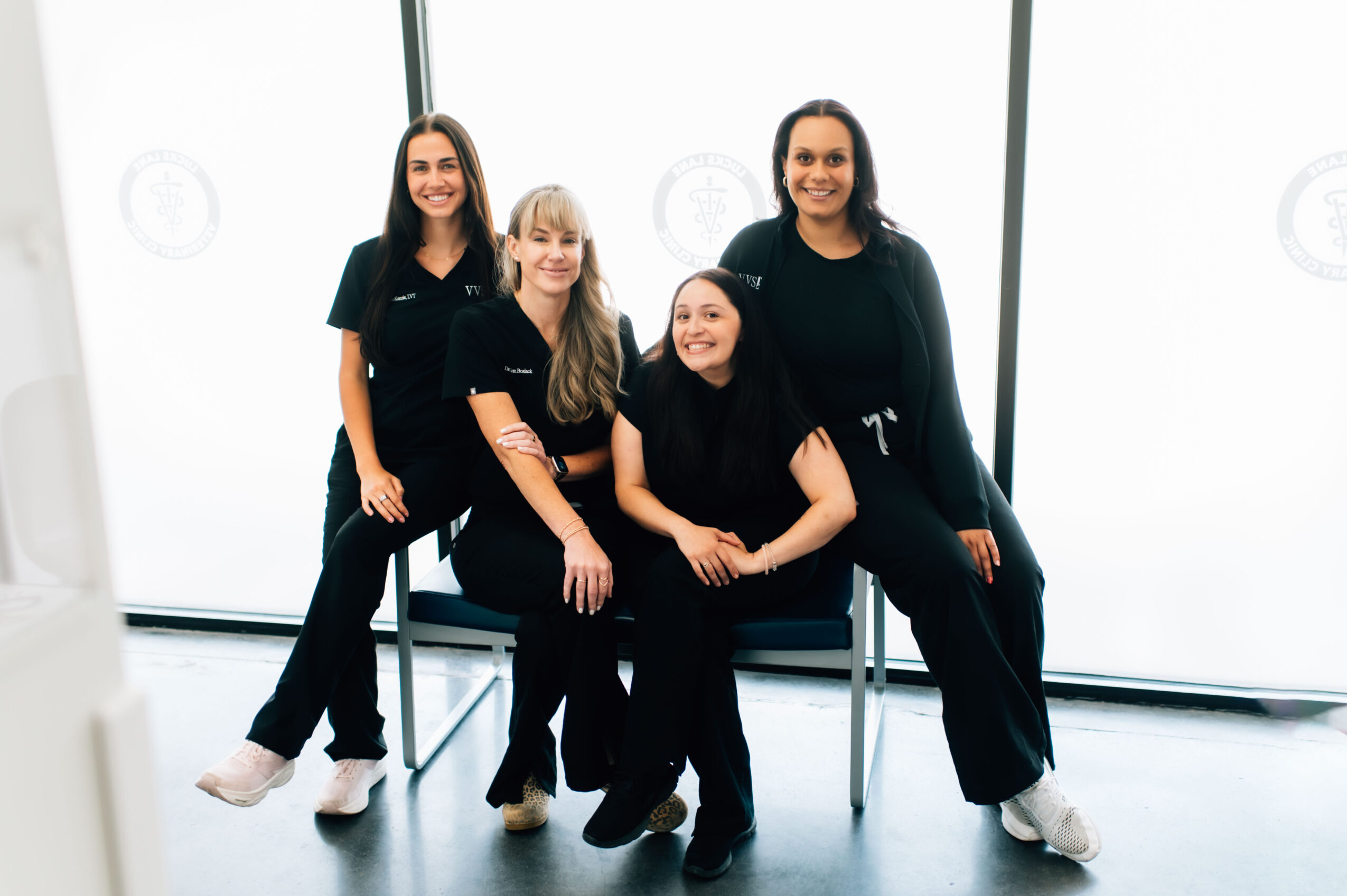 Four women sitting and standing in front of large windows, smiling, wearing black outfits, in a bright room.