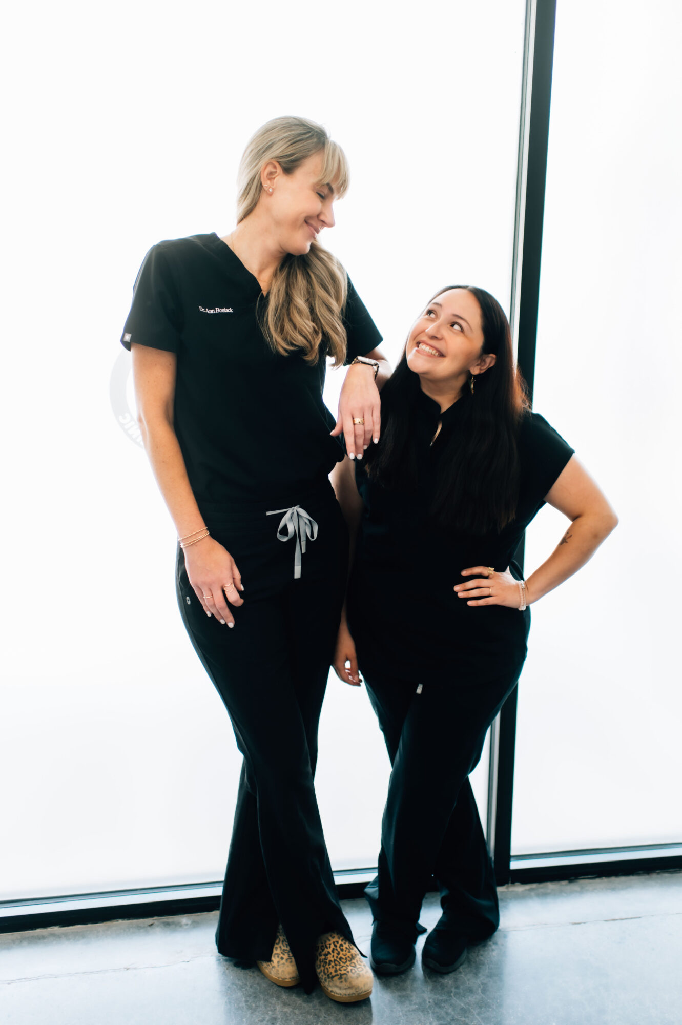 Two women in black uniforms smiling and talking indoors near large windows, one taller and the other shorter, with casual poses.