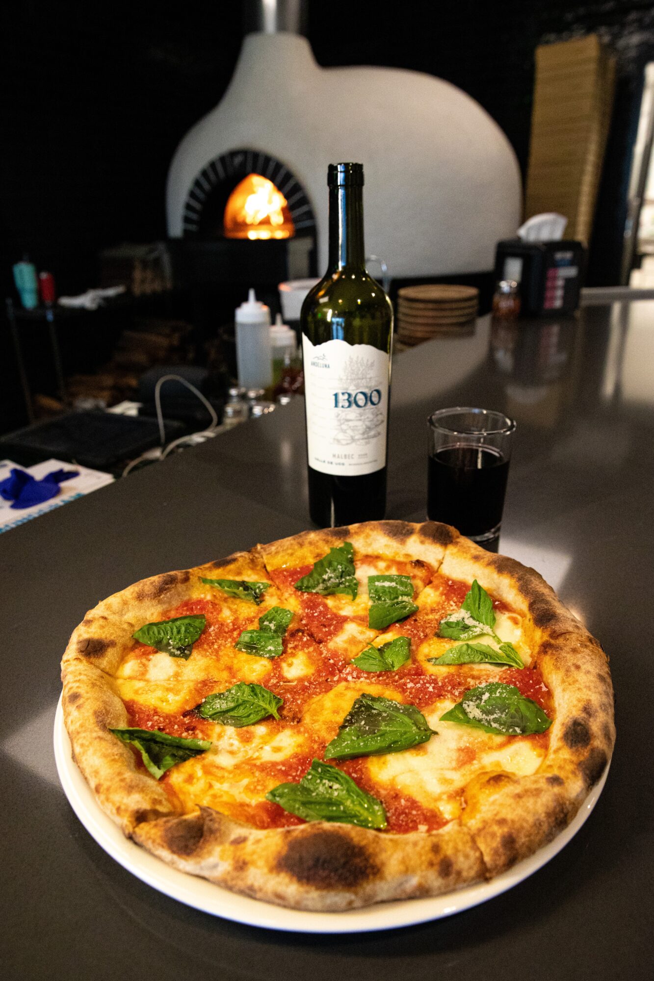 Pizza with green peppers and cheese on a white plate, bottle of wine, glass, and a fireplace in the background.