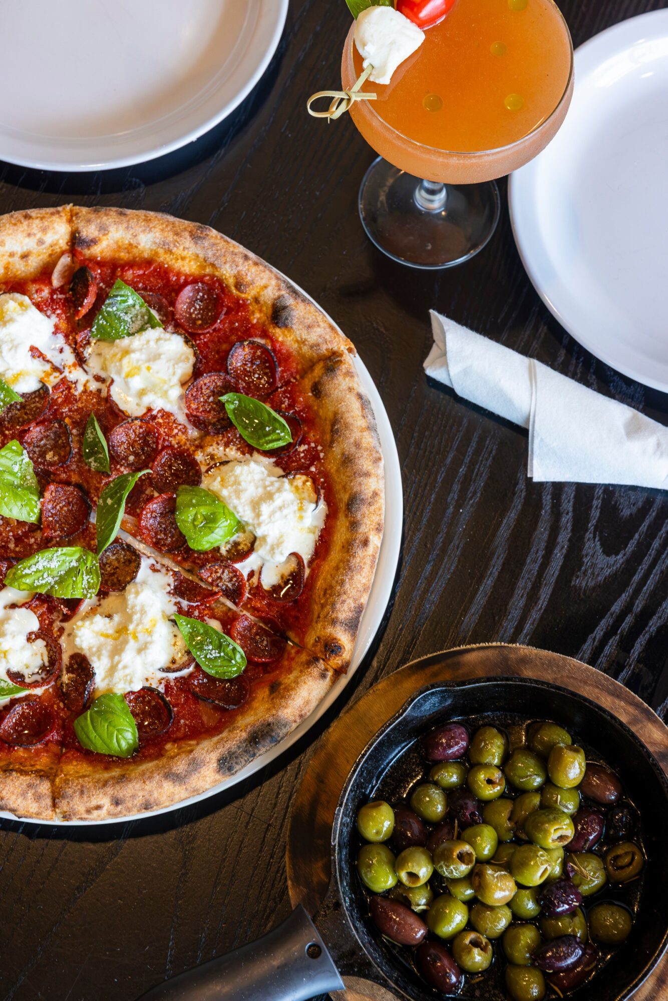 Pizza with basil, cheese, and tomato slices on a dark table, with a glass of drink and a bowl of green olives nearby.