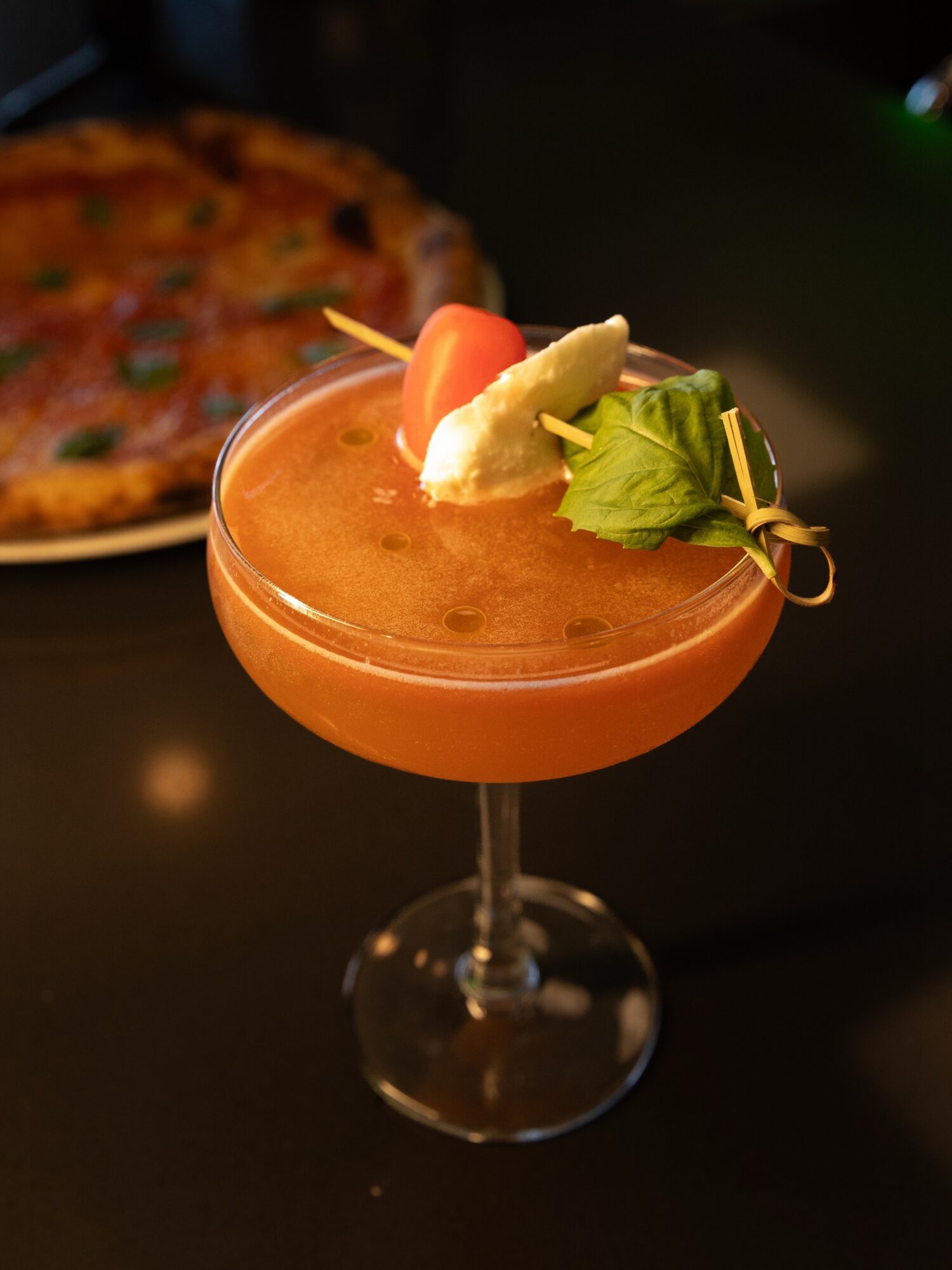 A cocktail garnished with a cherry, pineapple, and lettuce on a skewer, with a pizza in the background.