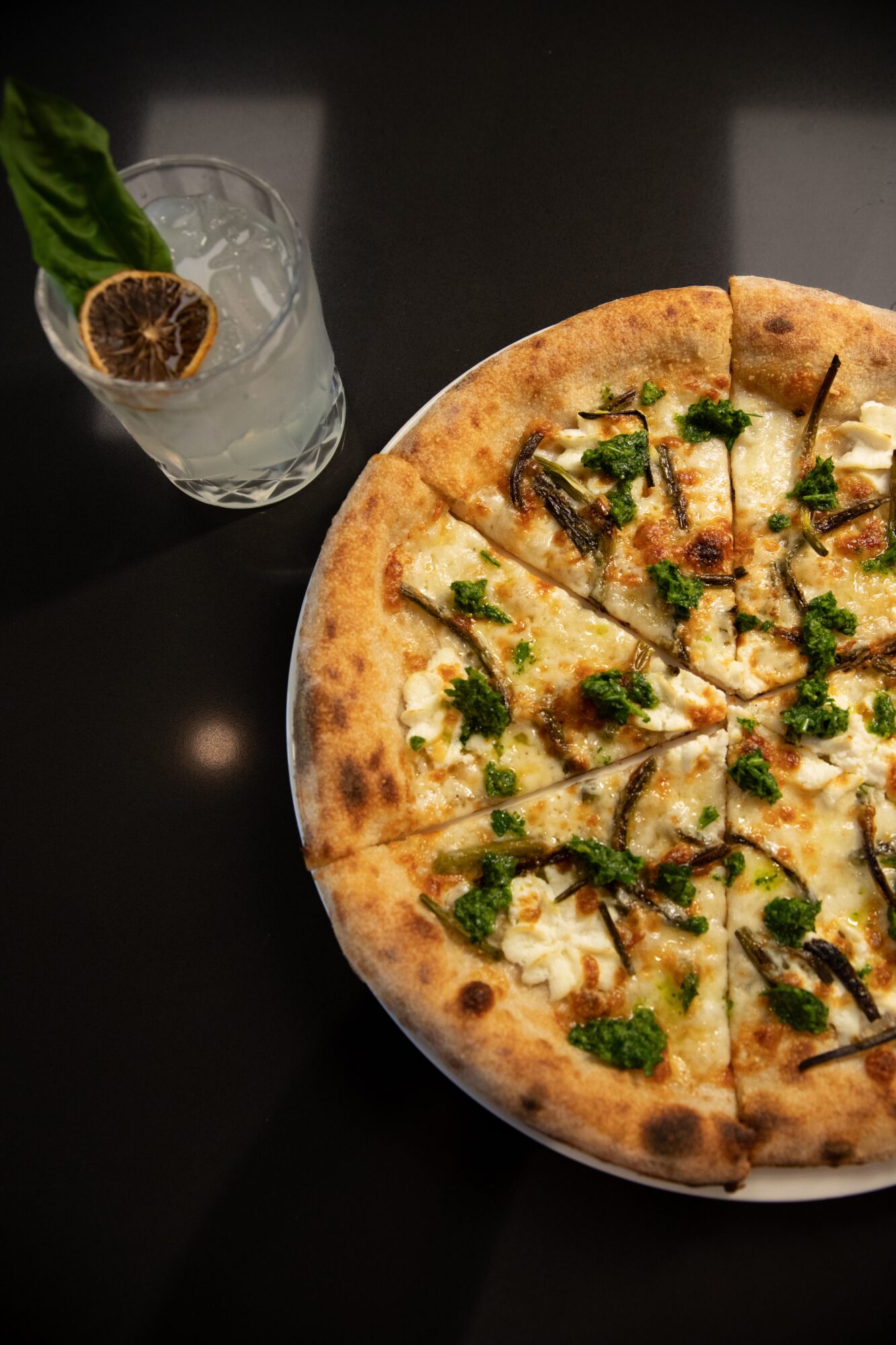 Pizza with toppings on a dark surface, alongside a glass with a lemon slice and green leaf, viewed from above.