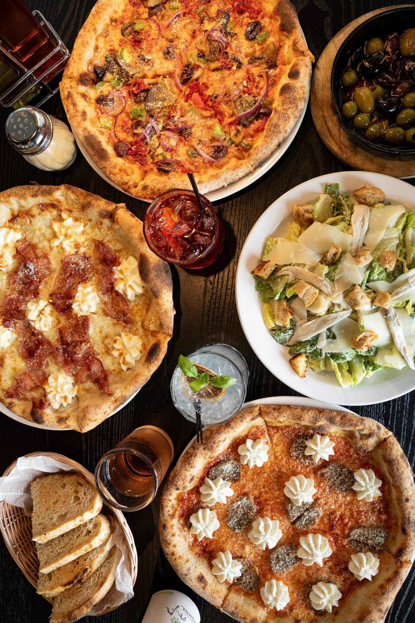 Pizza, salad, bread, and drinks on a table, with a bowl of olives and a glass of soda visible.