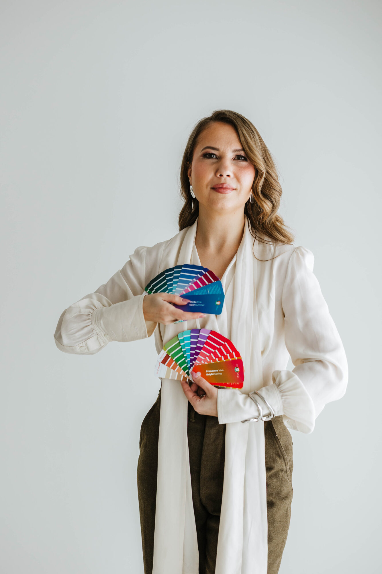 Woman with wavy hair holding colorful fan-shaped objects, wearing a white blazer and brown pants, standing against a plain light background.