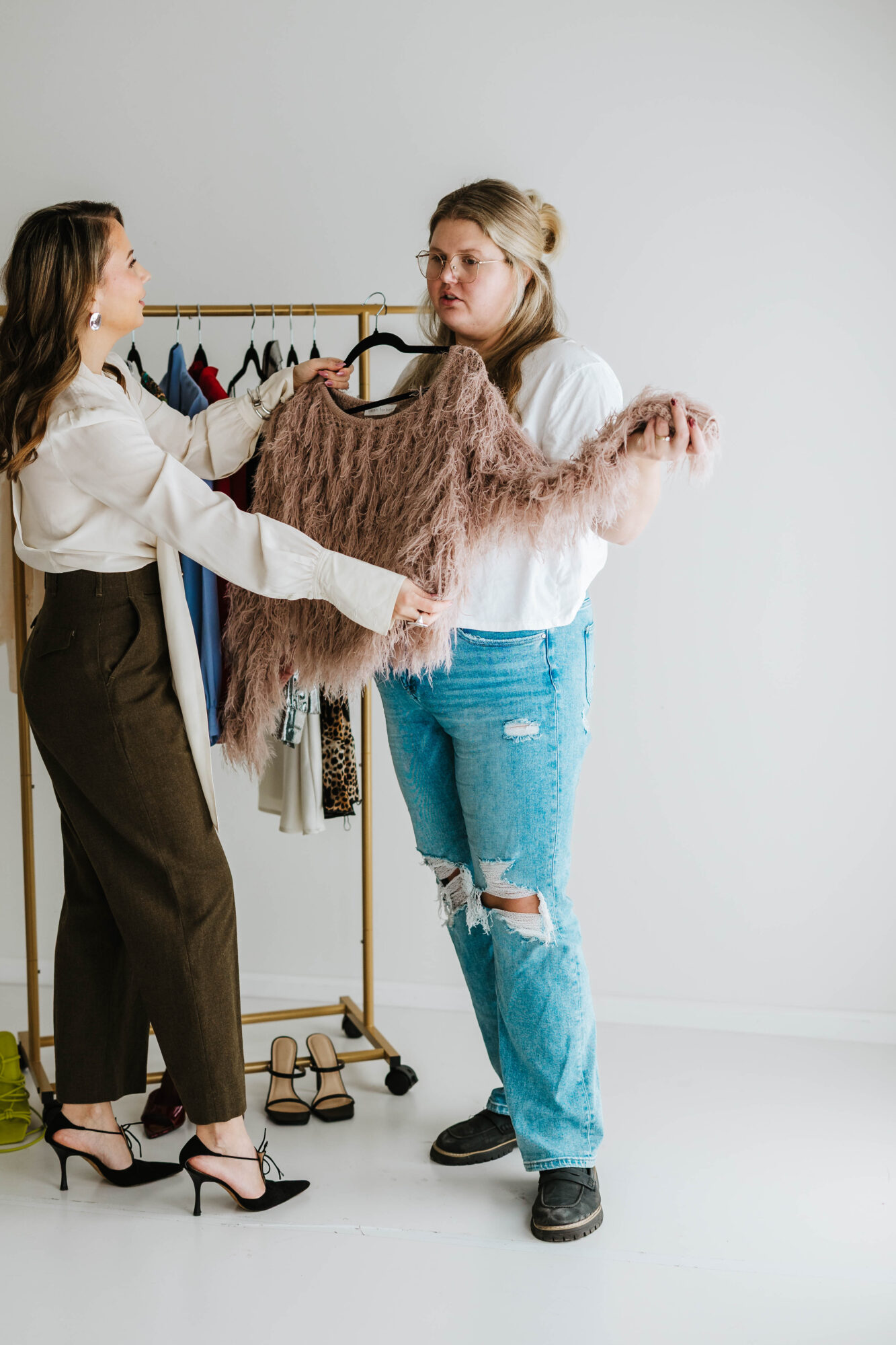 Two women exchange a sweater in a room with a clothing rack, one wearing black pants and heels, the other in ripped jeans and sneakers.