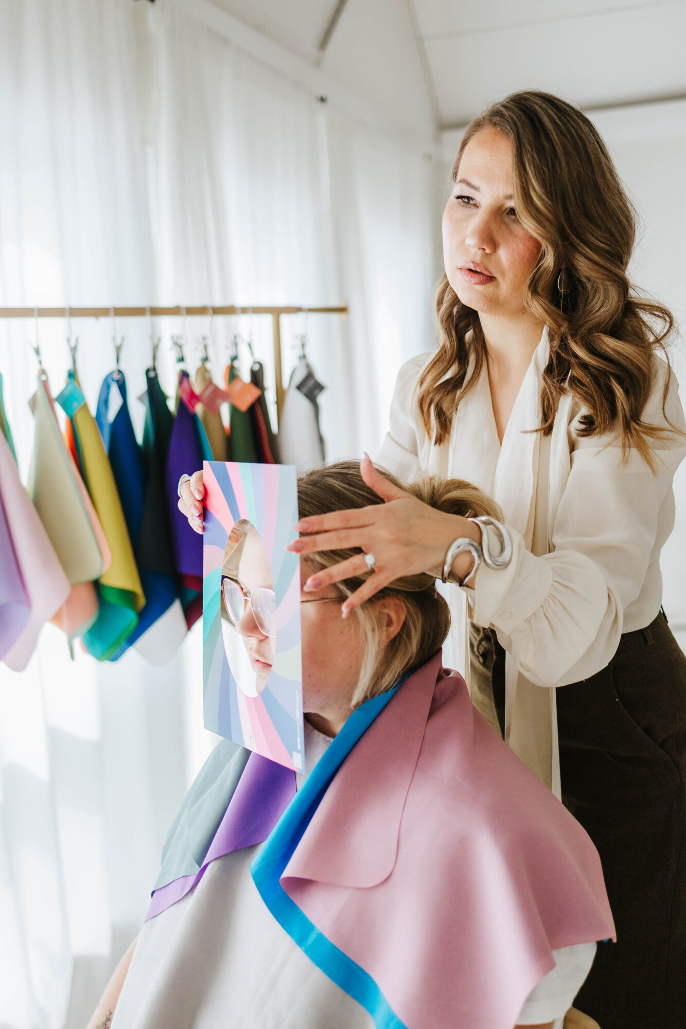 A woman with long wavy hair and a white blouse styles a child's hair in a salon with fabric swatches on the wall.