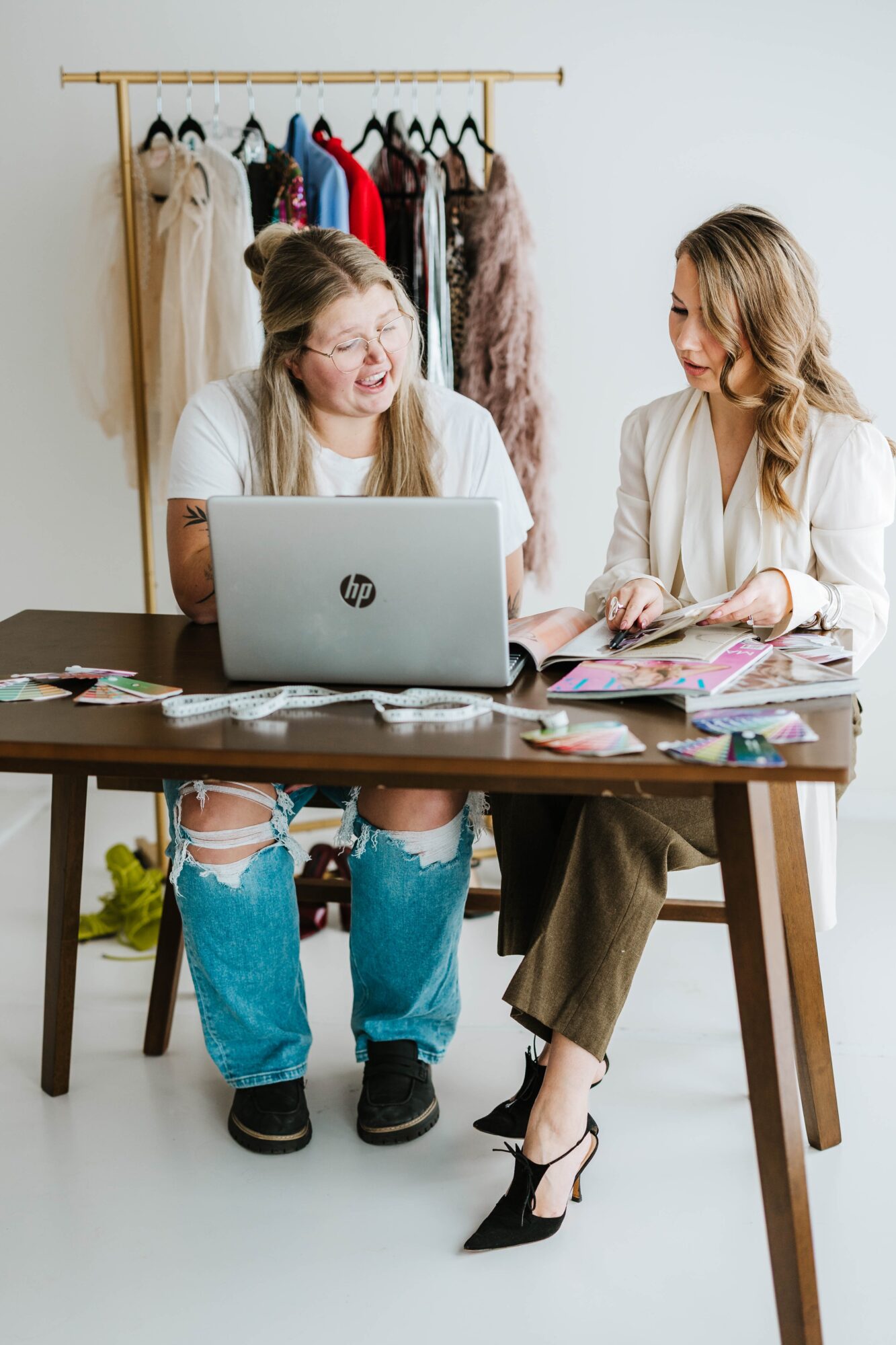 Two women sit at a table with a laptop, surrounded by clothing and papers, smiling and talking.