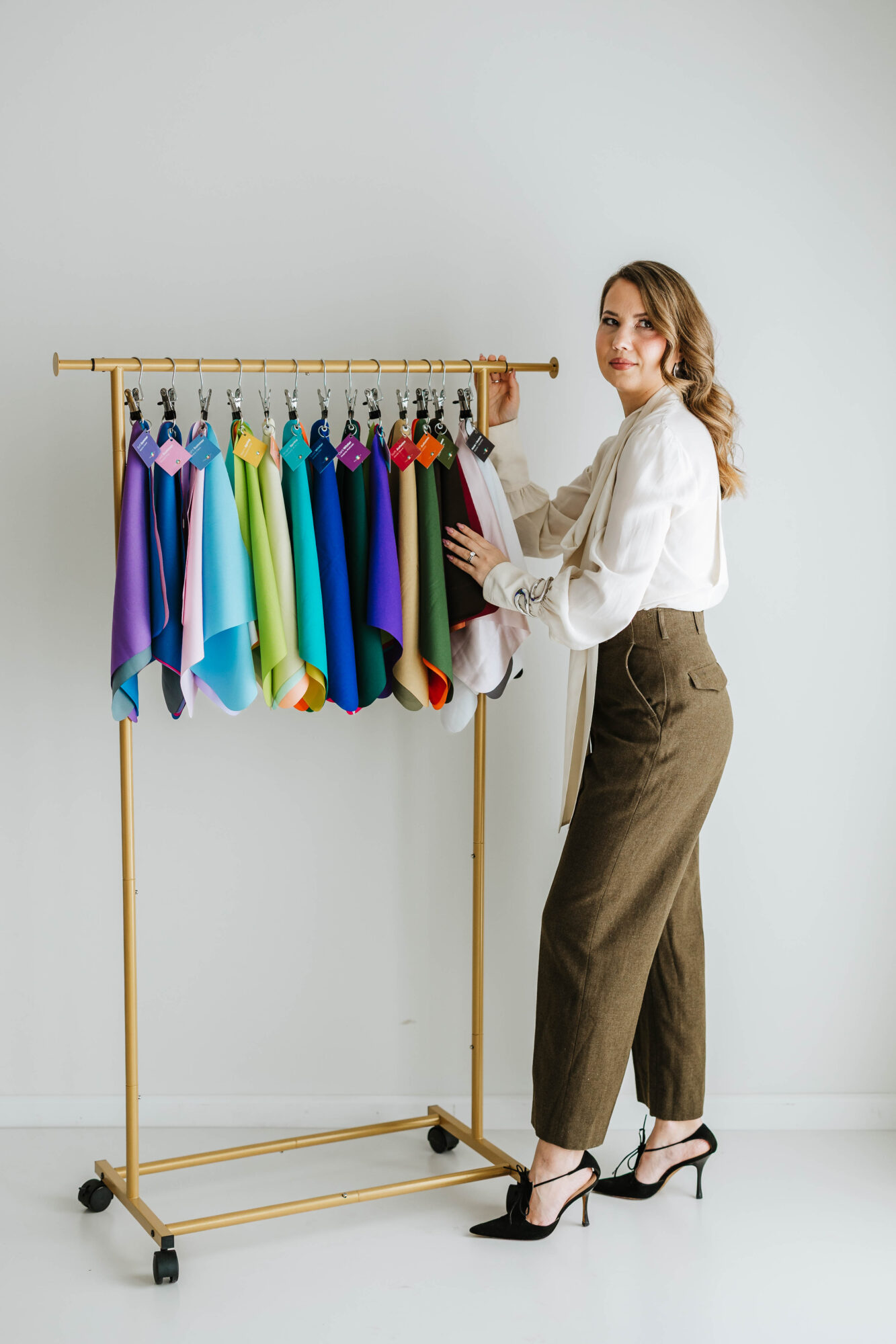 Woman with long hair in white blouse and brown pants stands next to a clothing rack with colorful garments, holding a book.