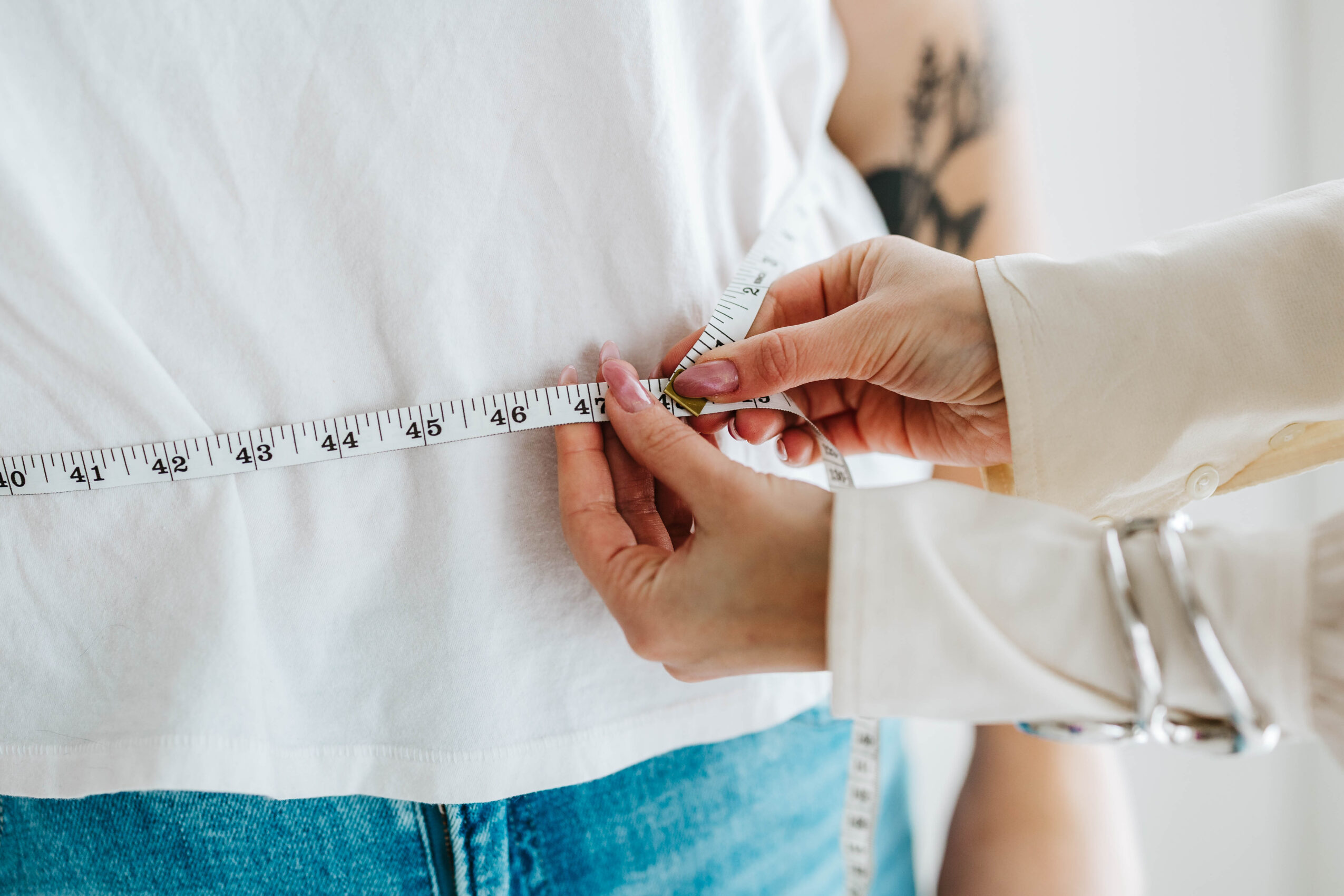 Person measuring another person's waist with a tape measure, wearing a white shirt and blue jeans.