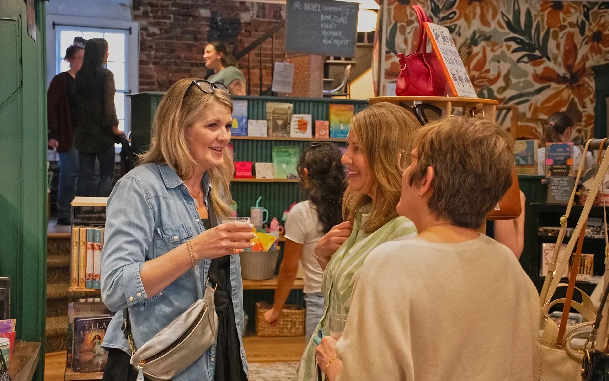 Two women and a young man are talking inside a bookstore, with books and a chalkboard in the background.