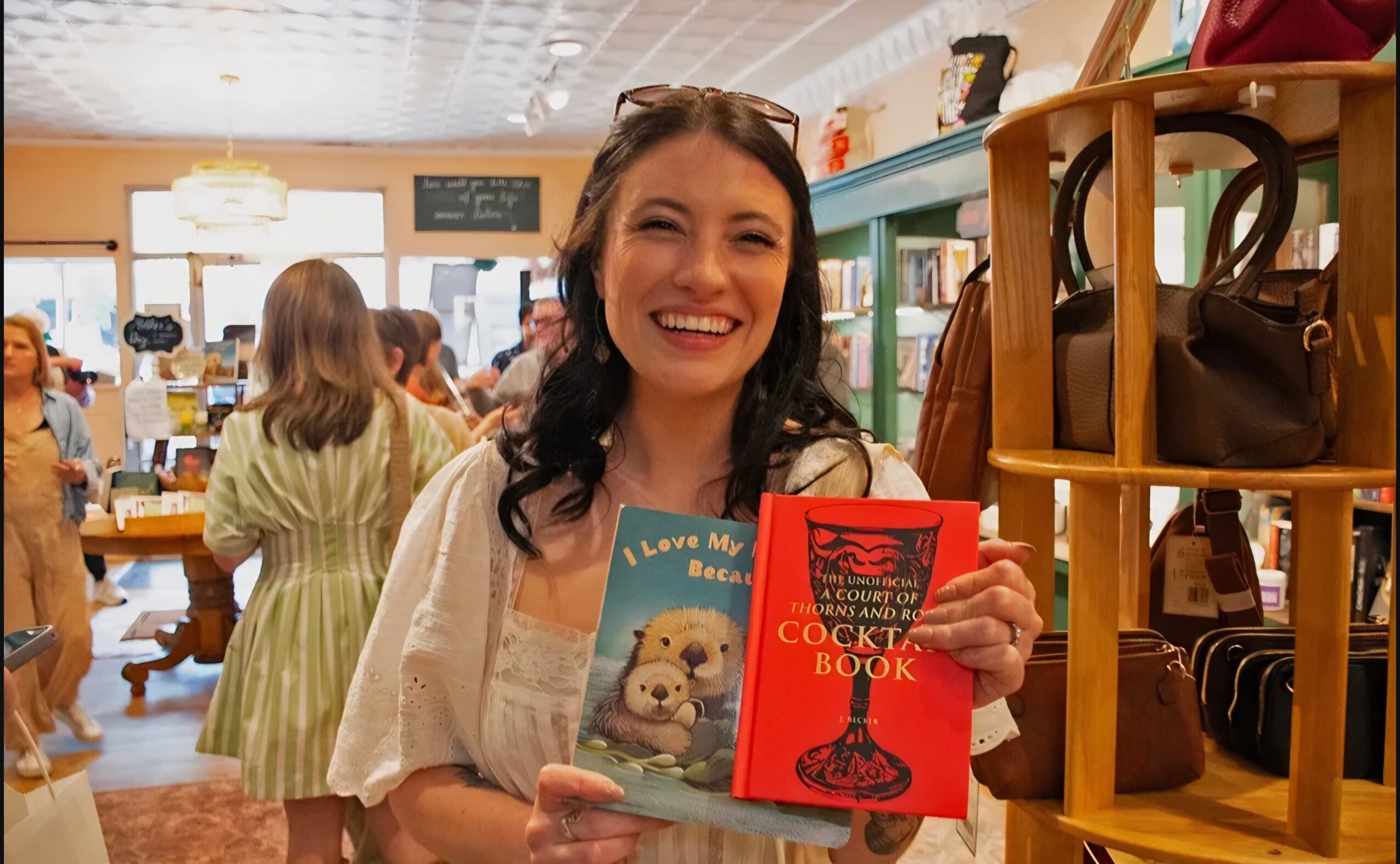 Woman smiling in bookstore holding a book and a red cocktail recipe book, shelves and other people in background.