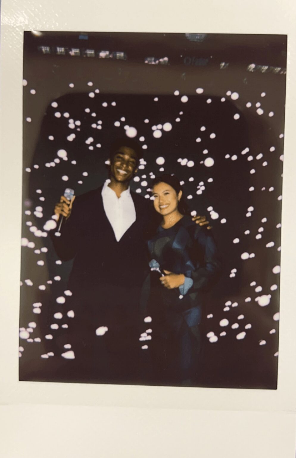Two people smiling, holding drinks, standing in front of a dark background with white dots, possibly at a celebration or event.