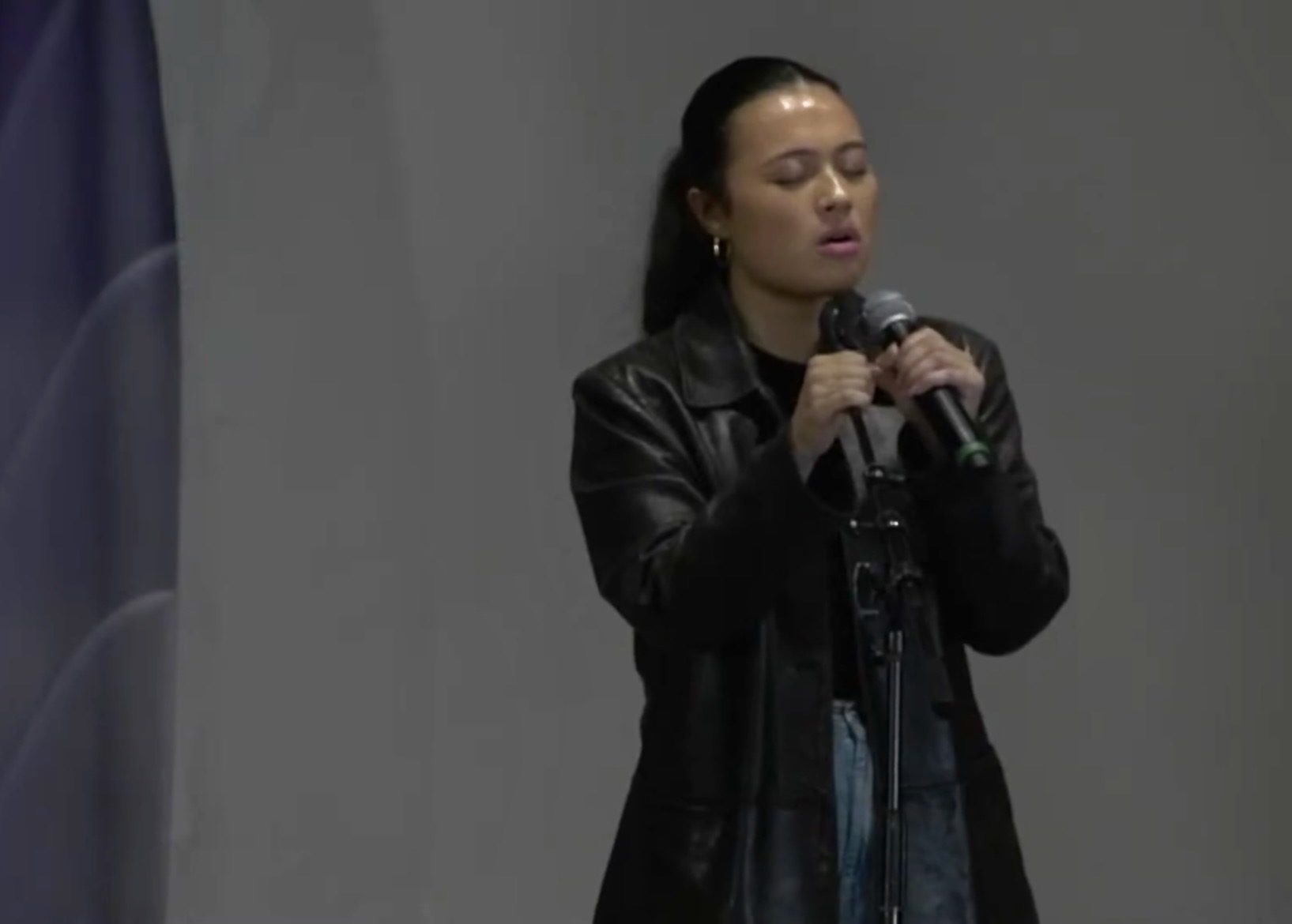 Woman with dark hair in a ponytail, wearing a black leather jacket, holding a microphone, standing against a plain background.