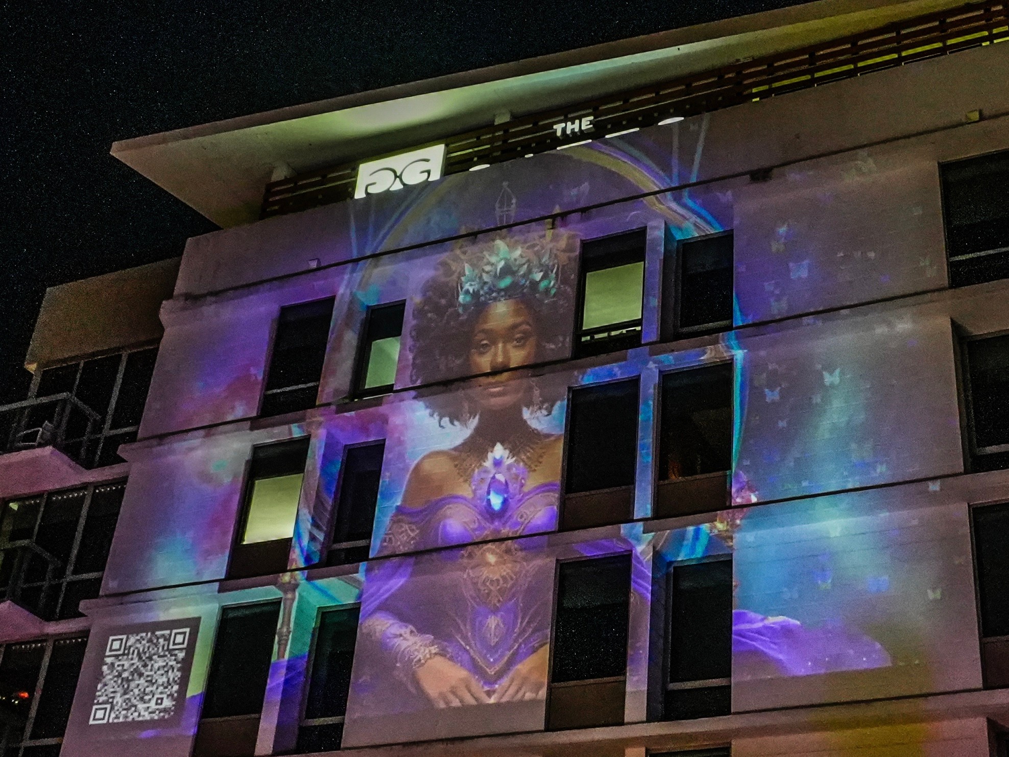 Projection of a woman with curly hair on a building at night, with colorful light effects and a QR code, 20 words.