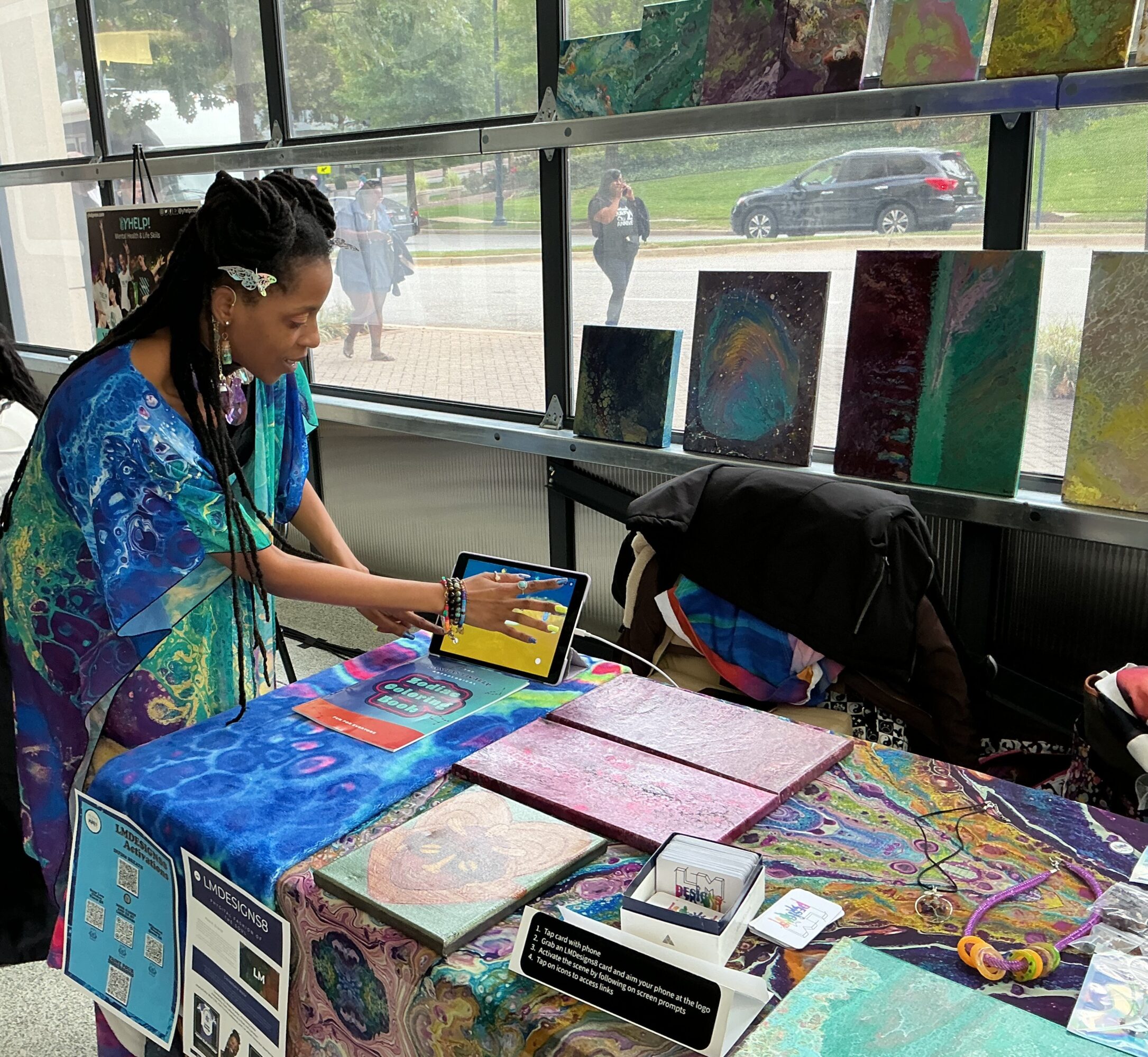 Woman in colorful dress looks at tablet at art display table inside a gallery, with paintings and informational signs visible.