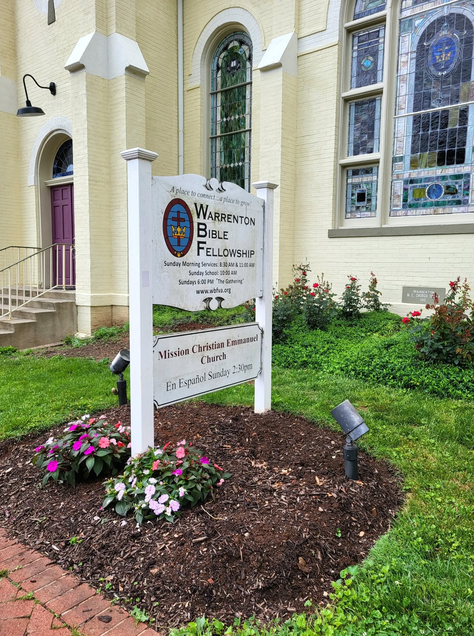 Church building with stained glass windows and a sign for Warren Baptist Bible Fellowship, surrounded by flowers and greenery.