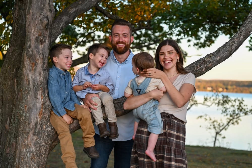 Family of five outdoors near a tree, smiling, with a lake in the background.