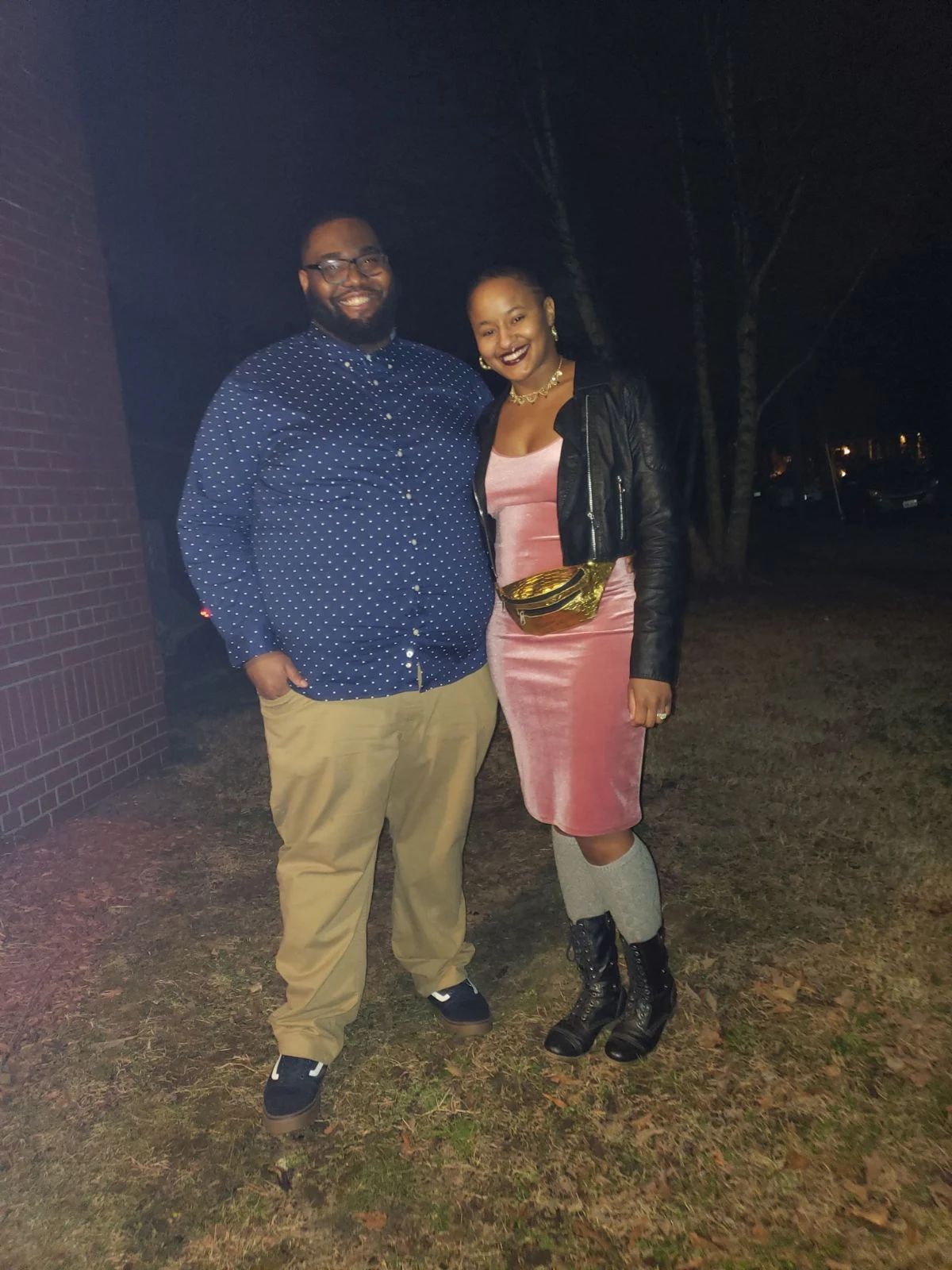 Two people standing outdoors at night, smiling, with a brick wall and trees in the background.