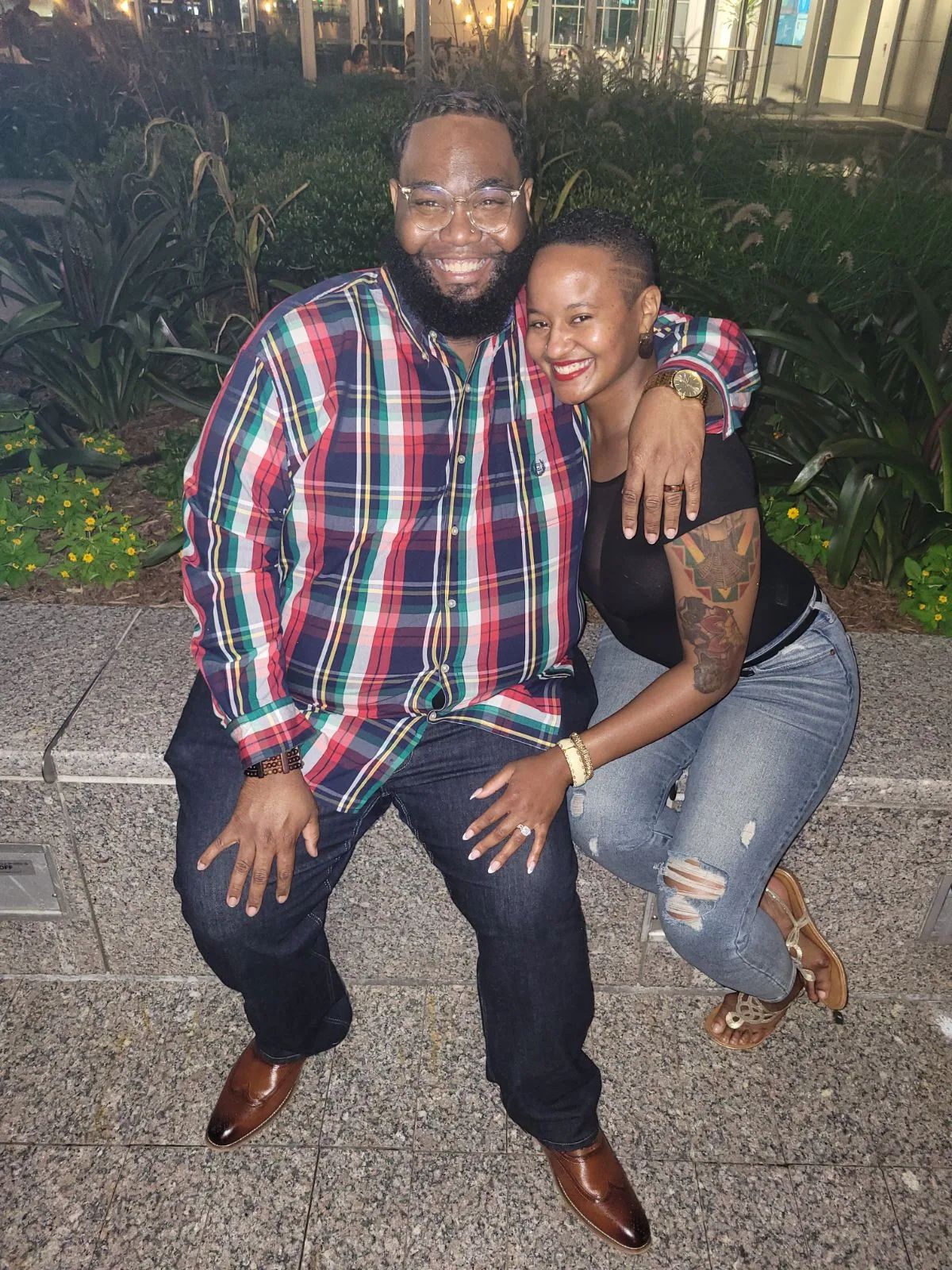 Two people sitting on a concrete ledge outdoors, smiling, with greenery and buildings in the background.