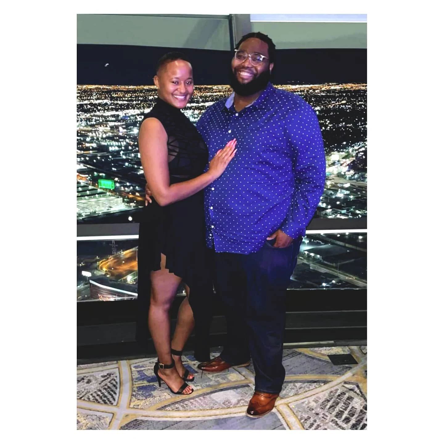 A smiling woman and man stand together indoors with a cityscape view behind them, woman in a black dress, man in a blue shirt.