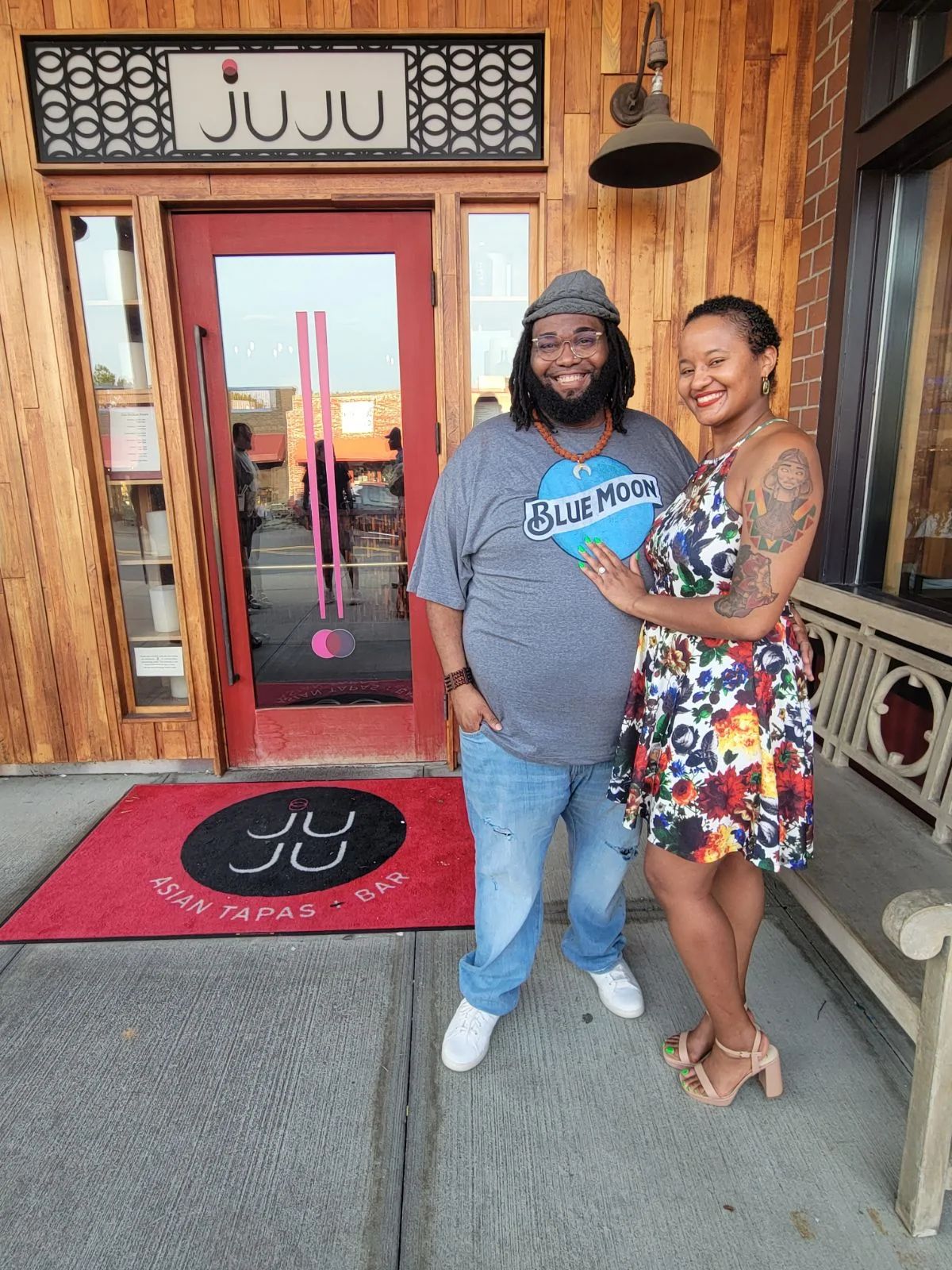 Two women stand outside a restaurant with wooden walls, smiling, one with a tattooed arm, the other wearing a blue shirt.