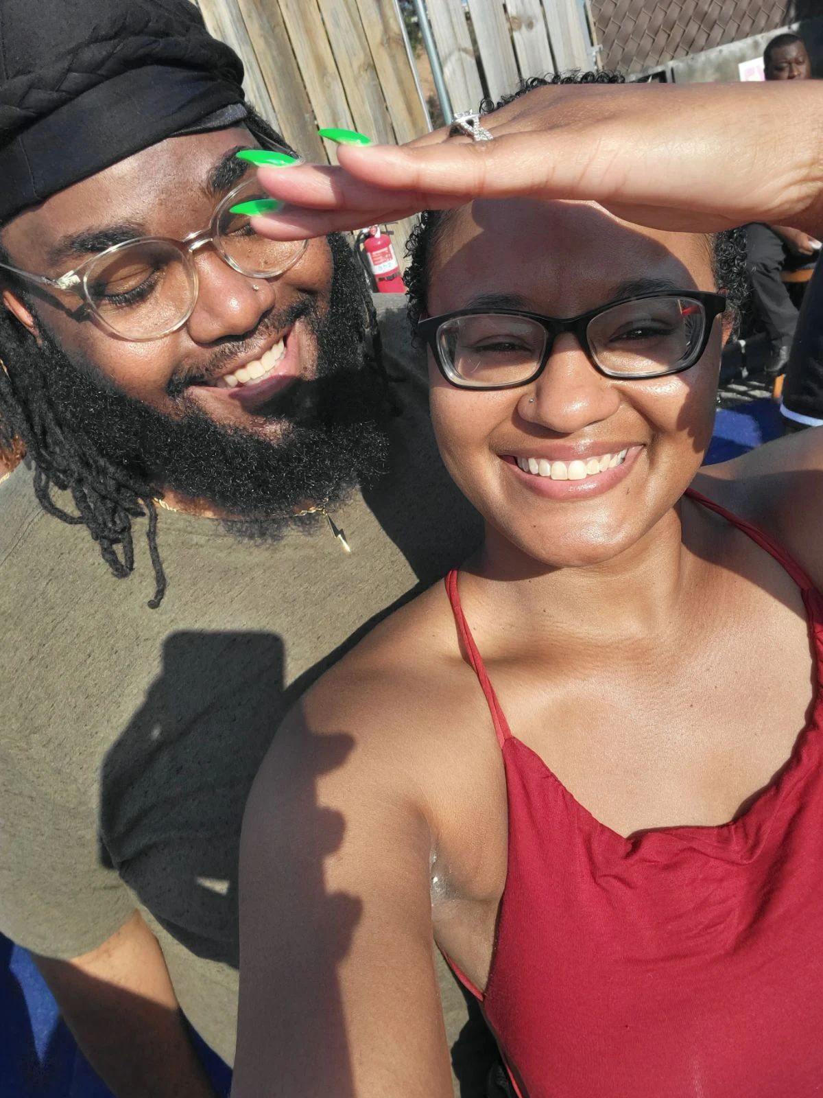 Two smiling people outdoors, one with glasses and a red top, the other with glasses and a black headwrap, saluting.