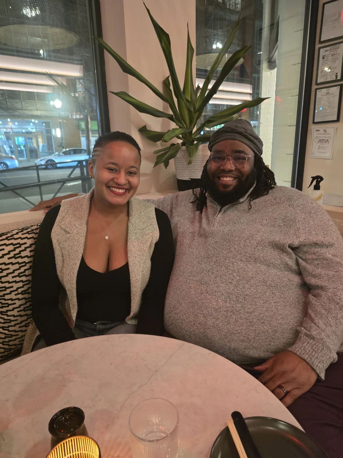 Two smiling people sitting at a table in a restaurant, with a large plant behind them and framed pictures on the wall.