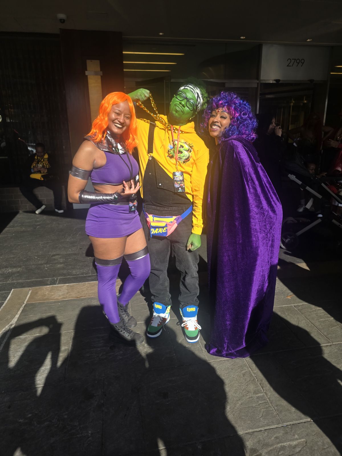 Three people standing together, two women in purple outfits and a person in a yellow jacket with balloons, smiling outdoors.