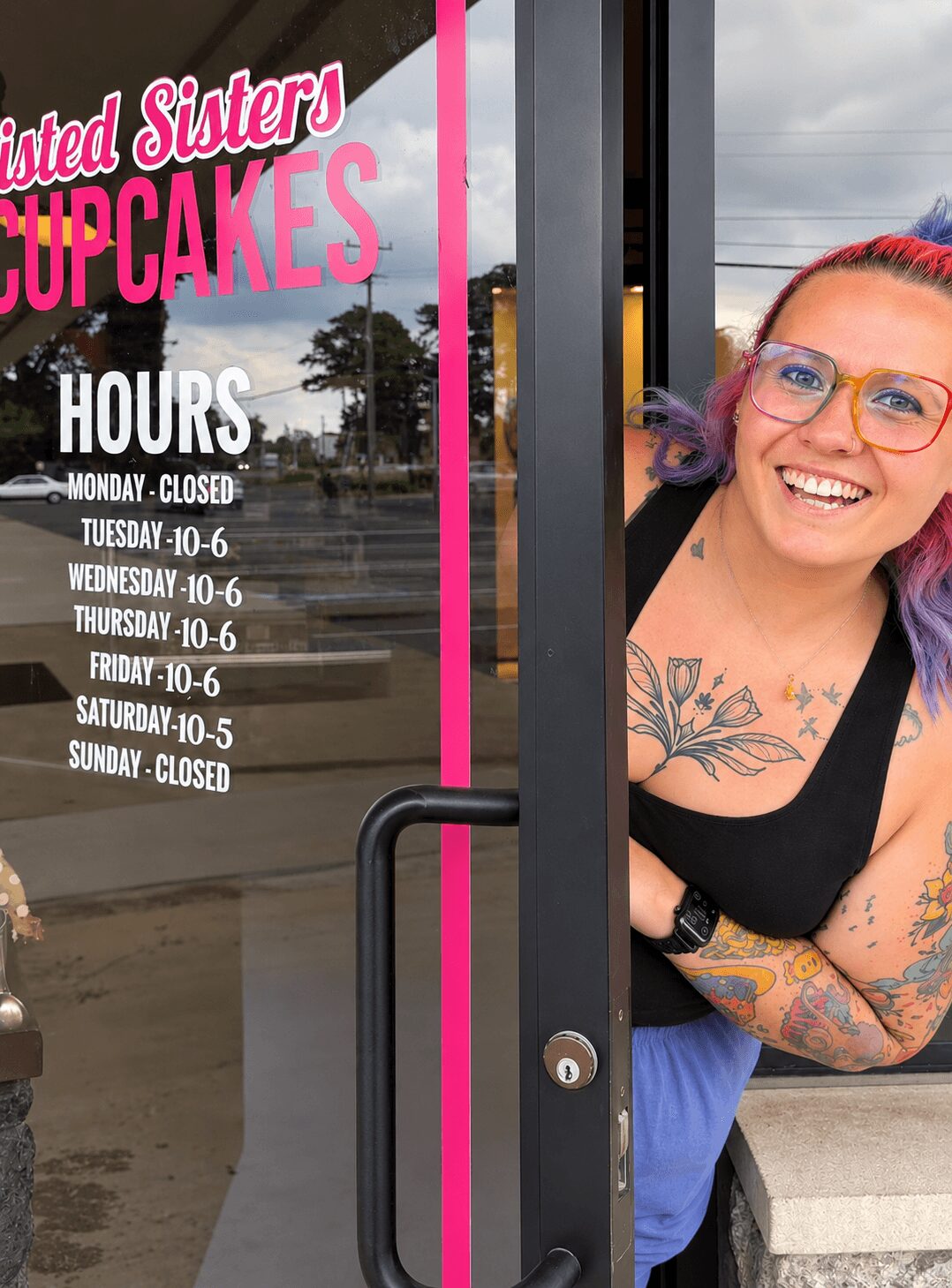 Smiling woman with colorful hair and tattoos peeks through door window, with cupcake shop hours on glass door.