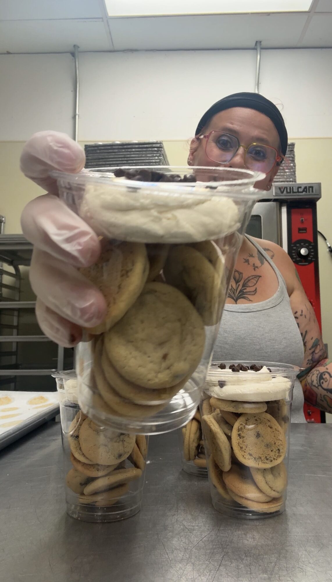Person holding a clear cup filled with cookies, with more cookies on the table, in a kitchen setting.
