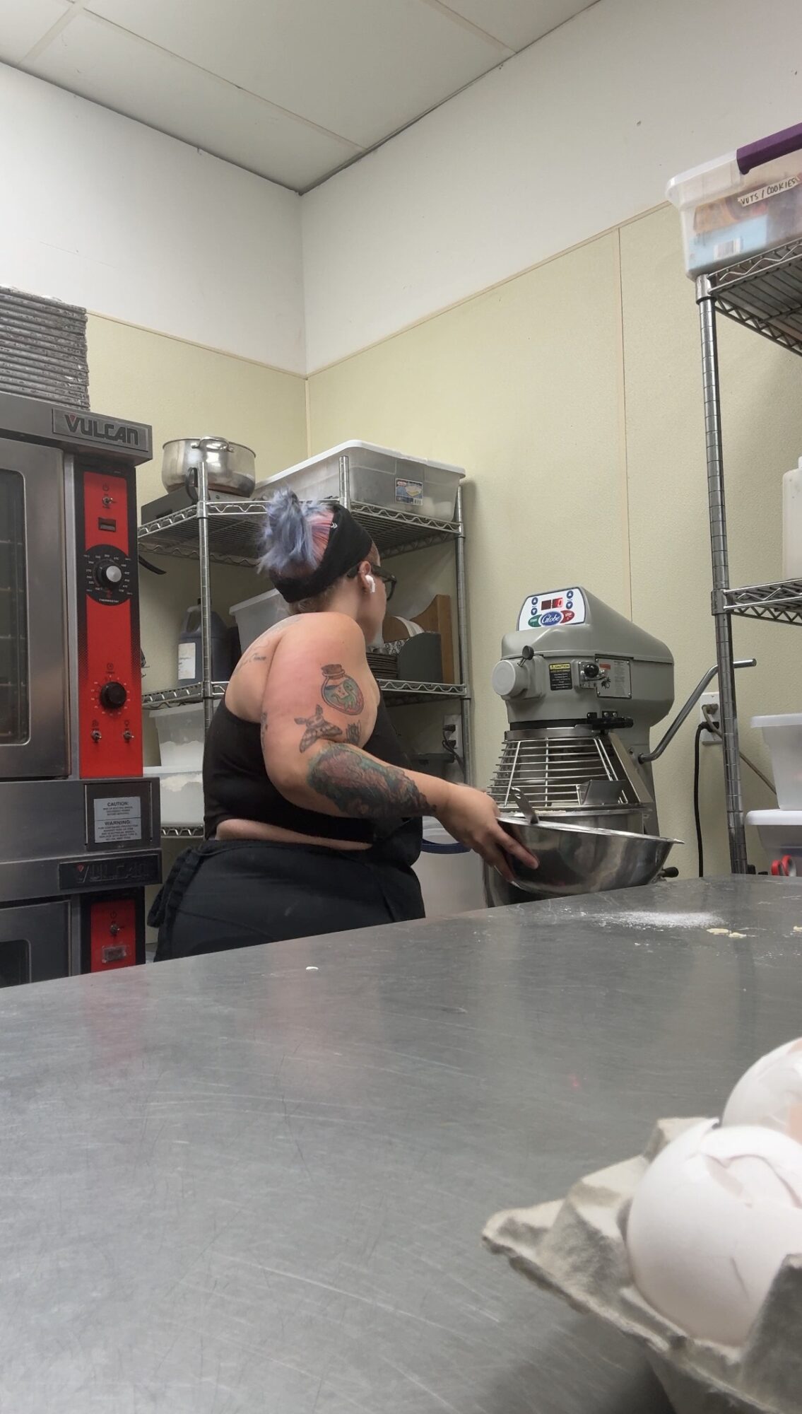 Person with tattoos wearing a hairnet and black shirt working in a kitchen with metal shelves and equipment.
