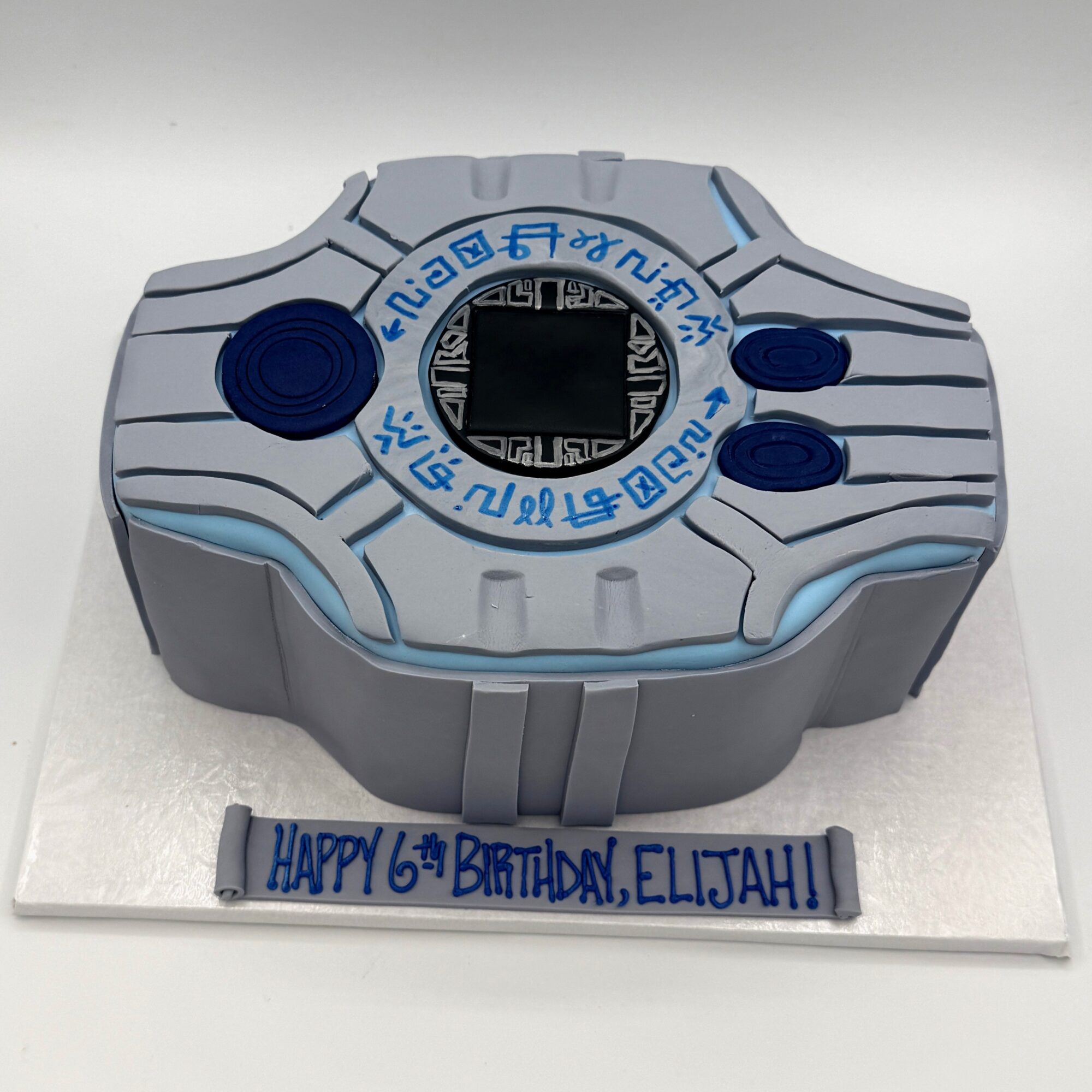 Digivice-shaped birthday cake with blue and gray colors, digital display, and a message reading 'HAPPY 6th BIRTHDAY, ELIZAH!'