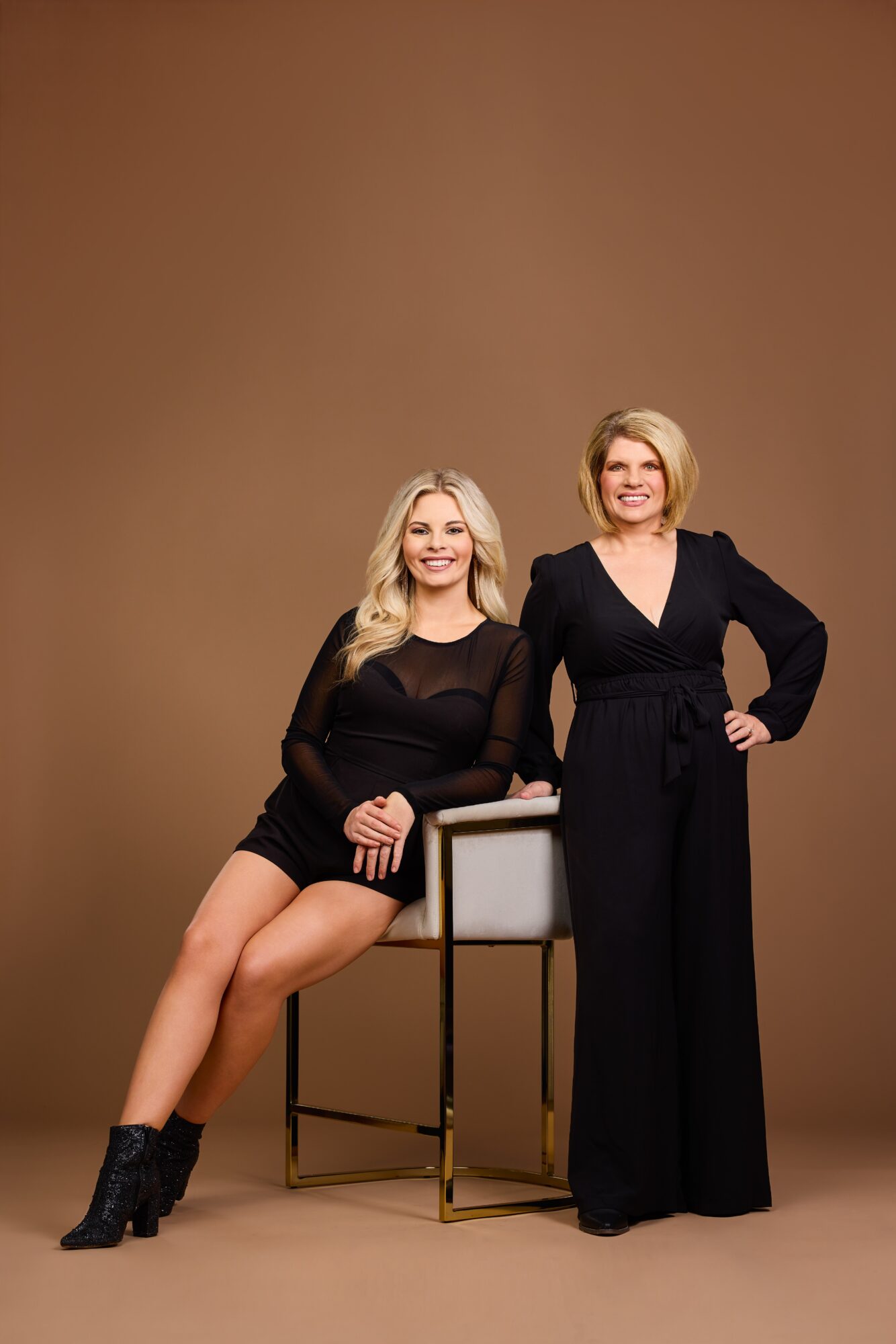 Two women pose together, one sitting on a chair and the other standing, against a plain brown background.