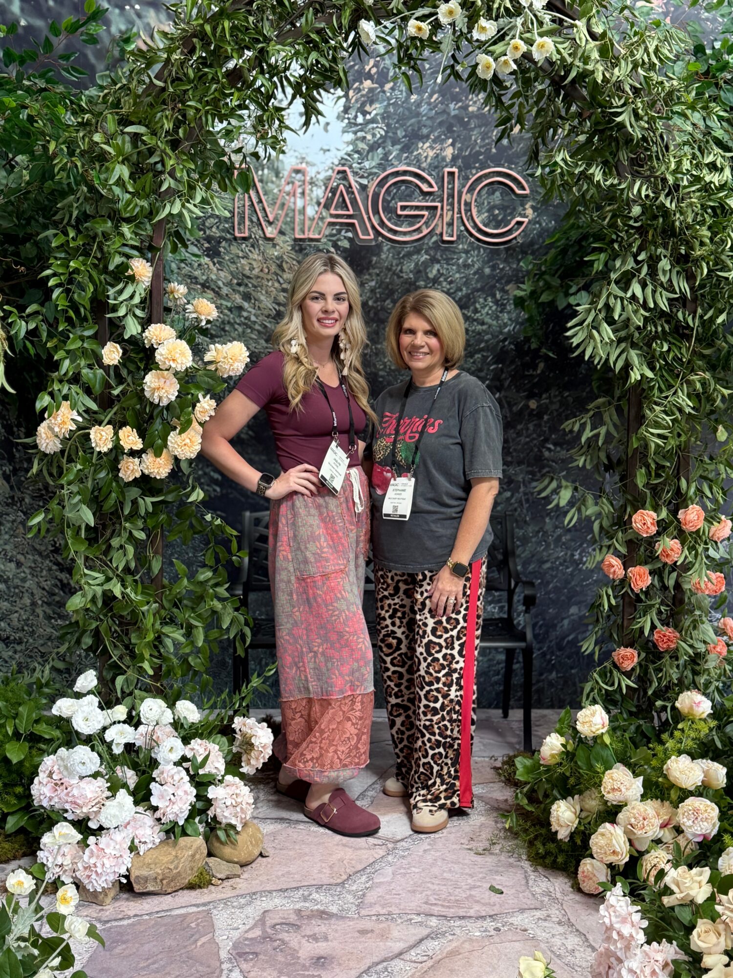 Two women stand in front of a floral arch with the word 'MAGIC' above them, surrounded by flowers and greenery.