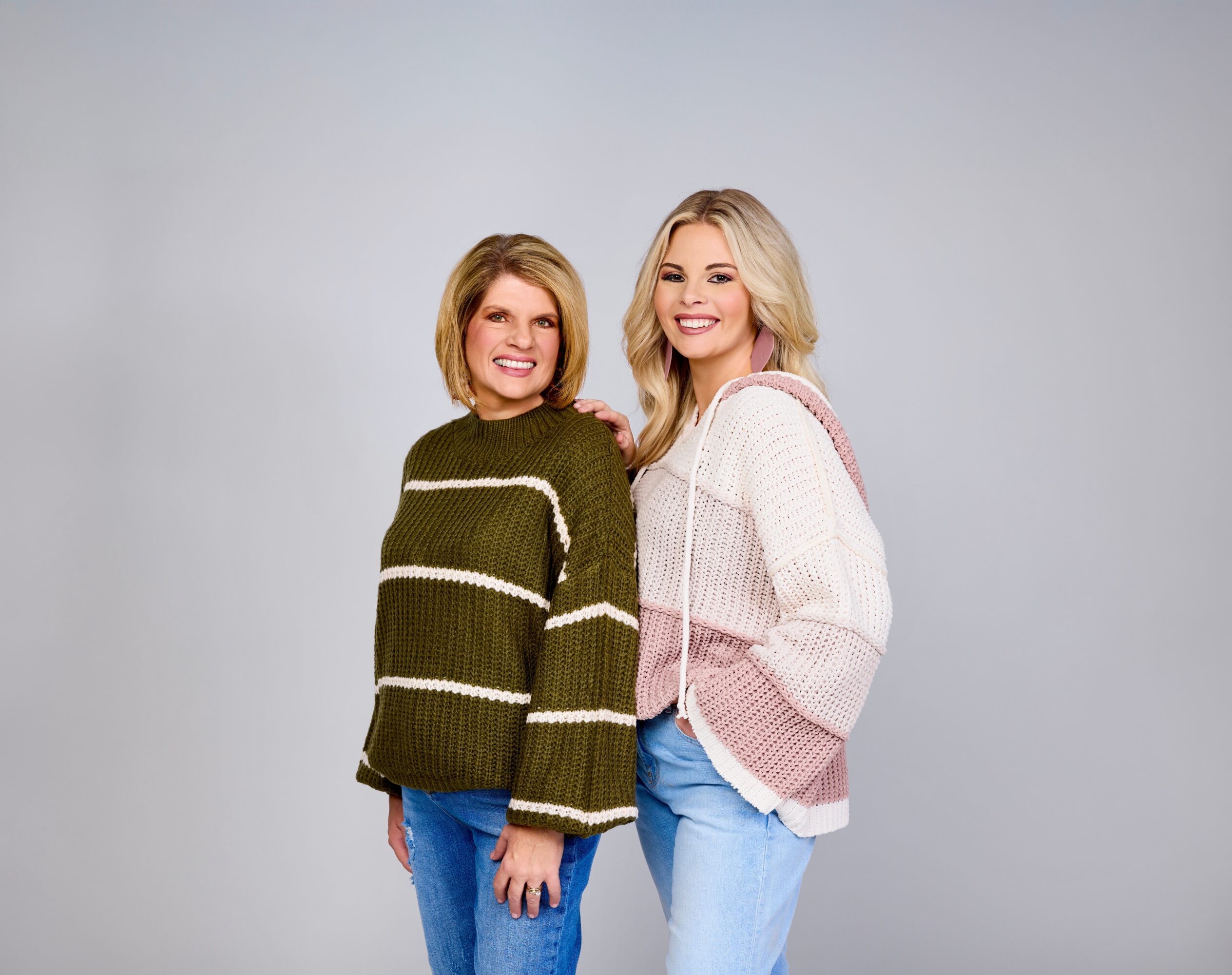 Two women stand side by side smiling, wearing casual sweaters and jeans, against a plain light background.