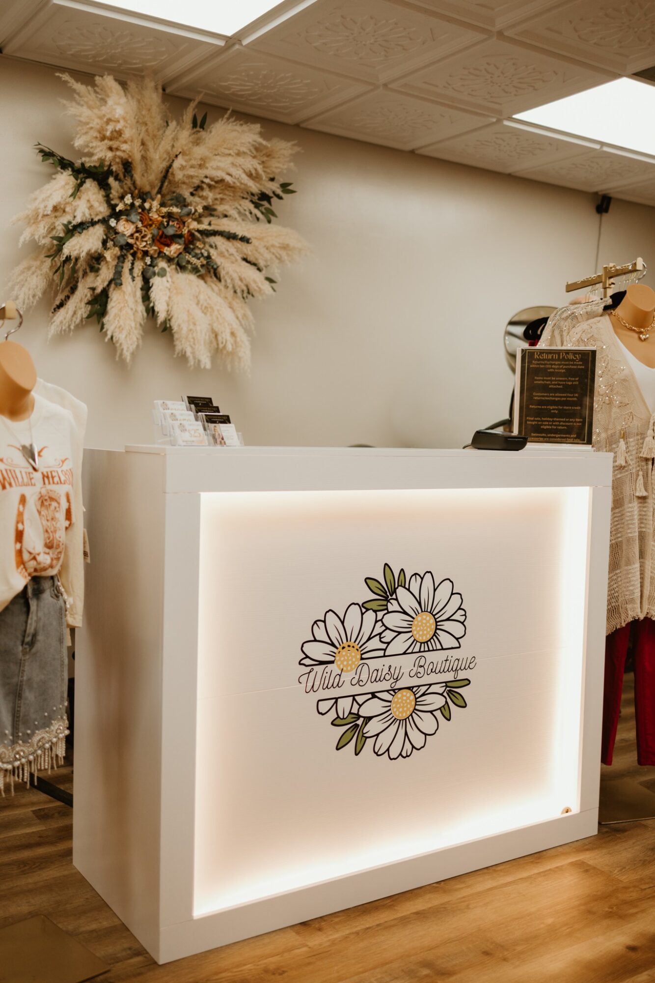 Reception desk with floral logo, large flower arrangement on wall, and a cash register, in a well-lit room.