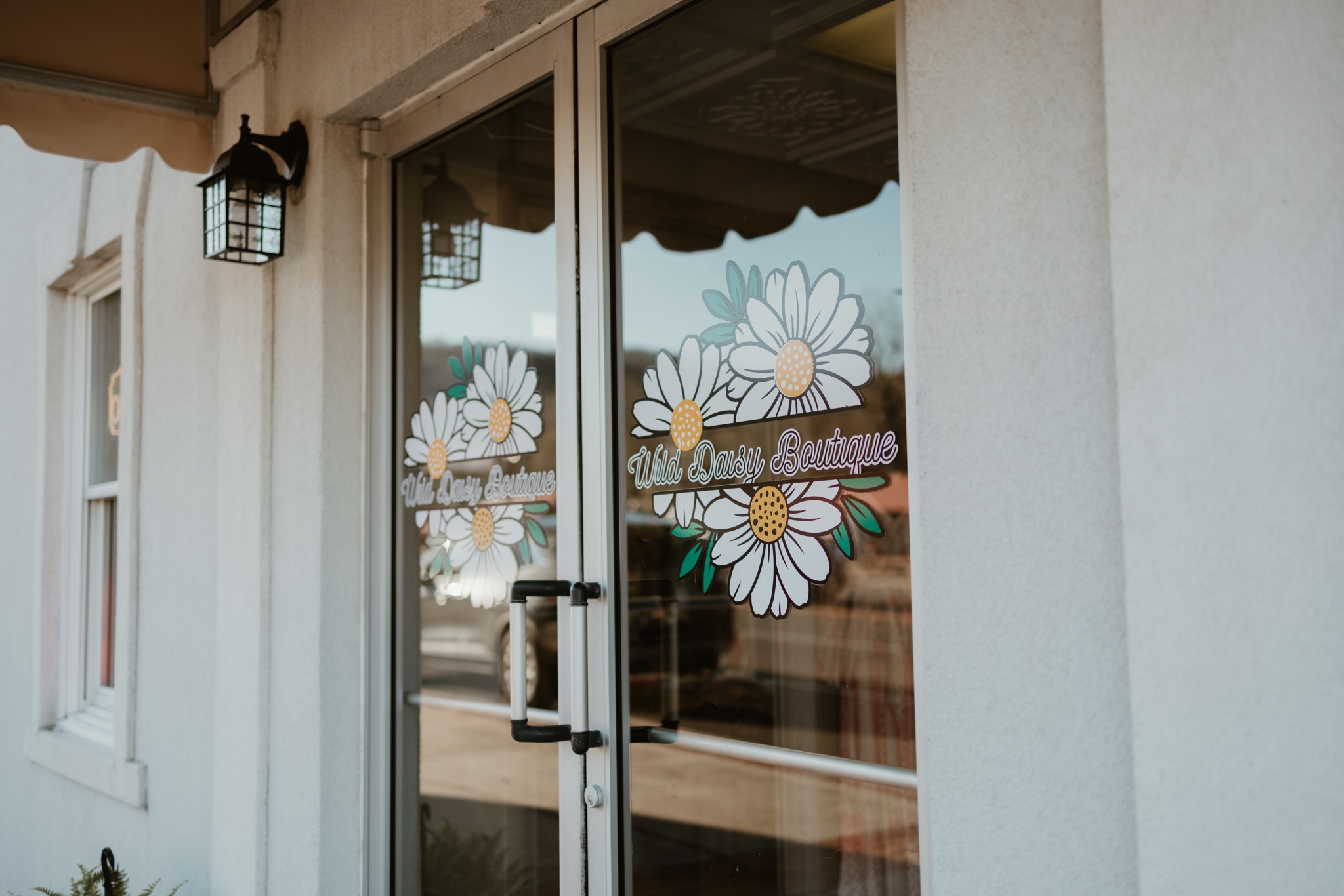 Glass door with daisy flower decals, white wall, and outdoor view, including a street and building, reflected in the glass.