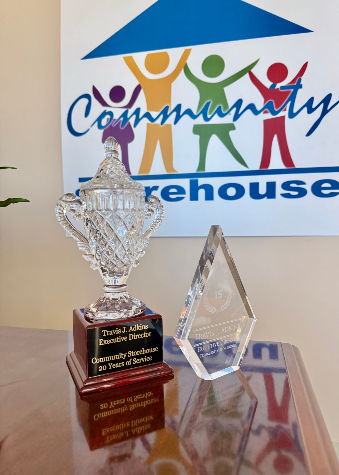 Trophy and glass award on a table in front of a colorful community logo with four stylized people holding hands.