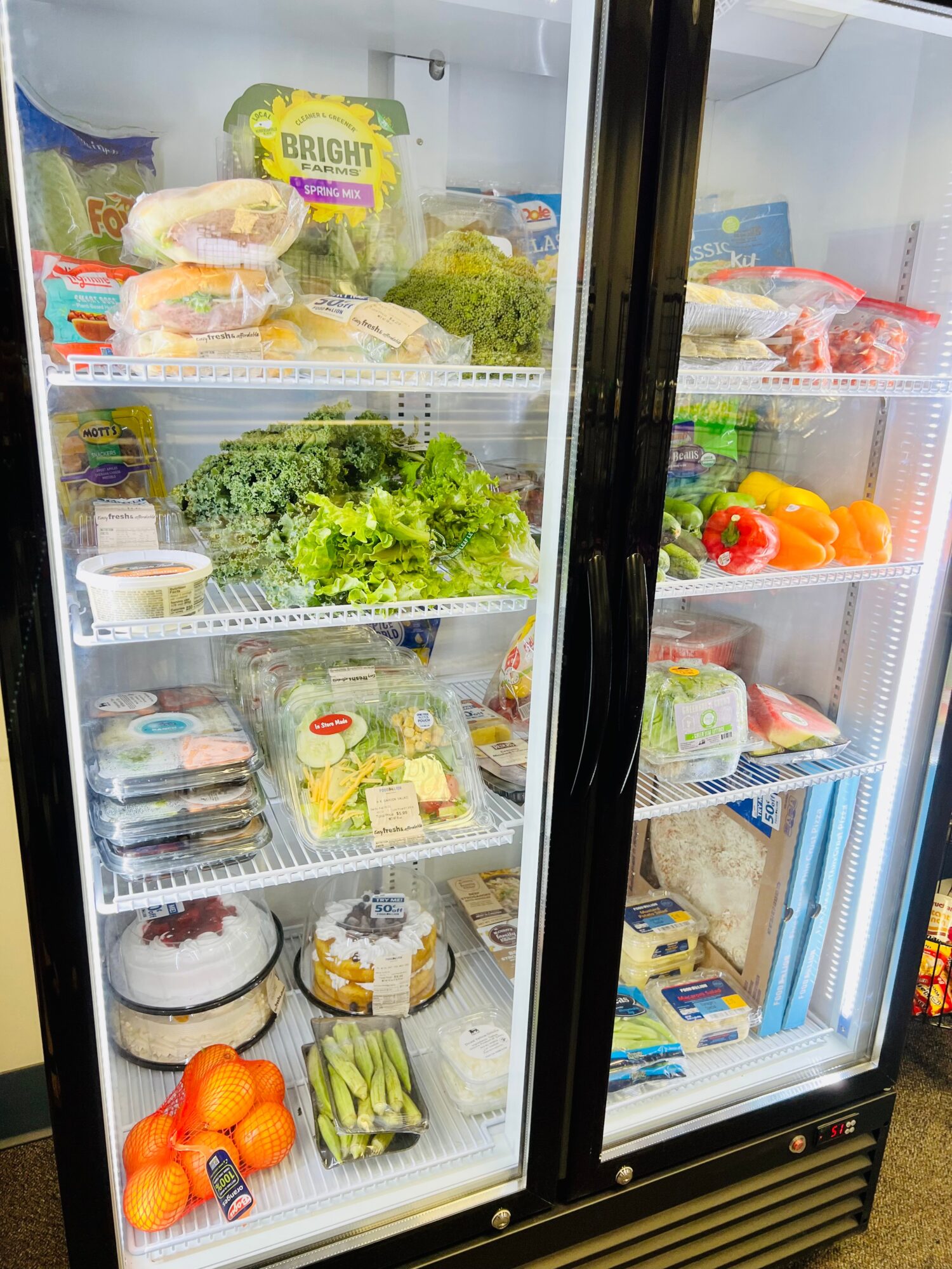 Refrigerator with vegetables, fruits, and packaged food items on shelves, door closed, in a grocery store or kitchen.