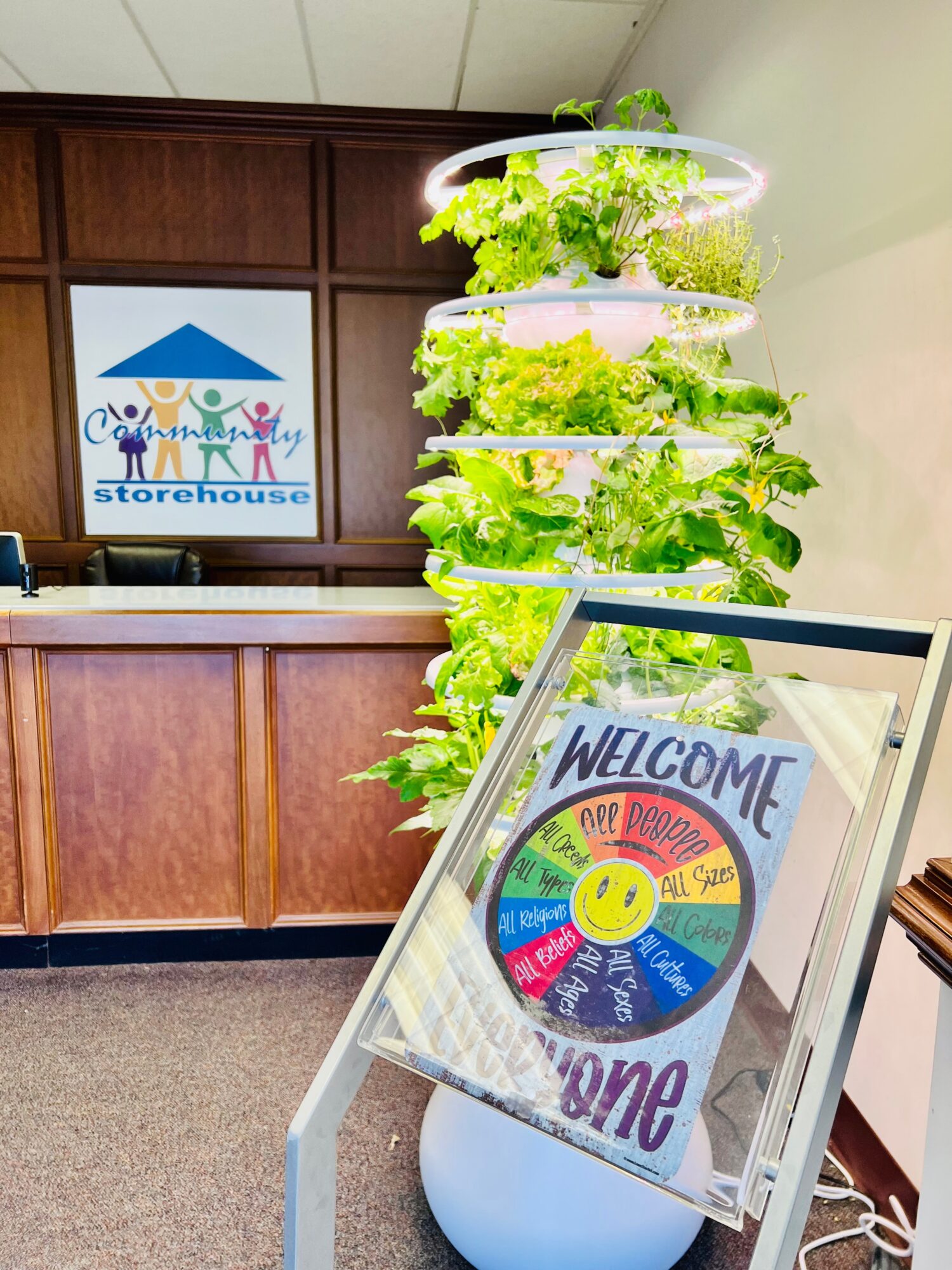 Indoor reception area with a vertical garden of green plants, a welcome sign with colorful text and a smiley face, and a wooden counter.